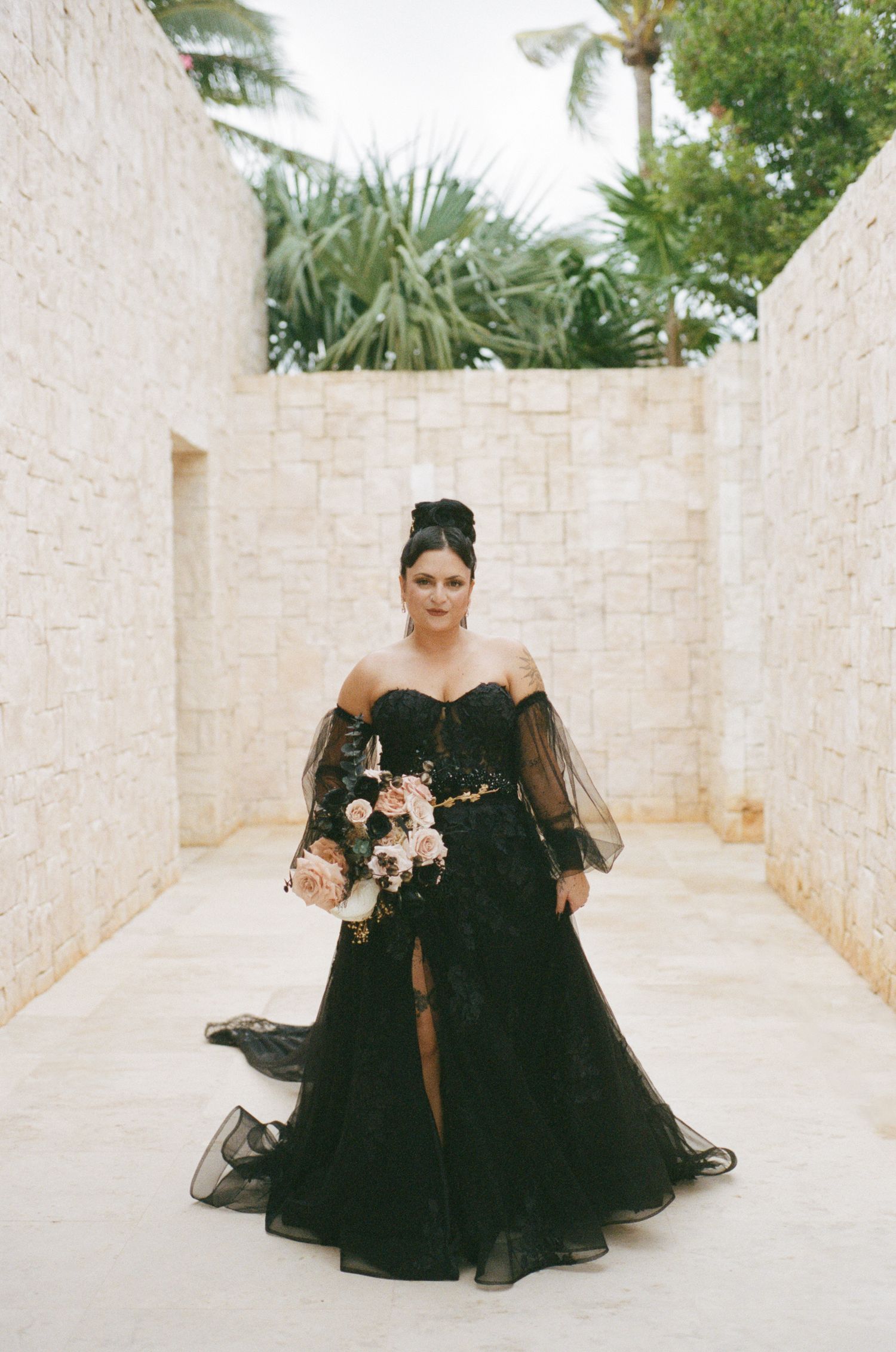 Bride in stunning black off-shoulder wedding gown with tulle skirt holding pink floral bouquet.