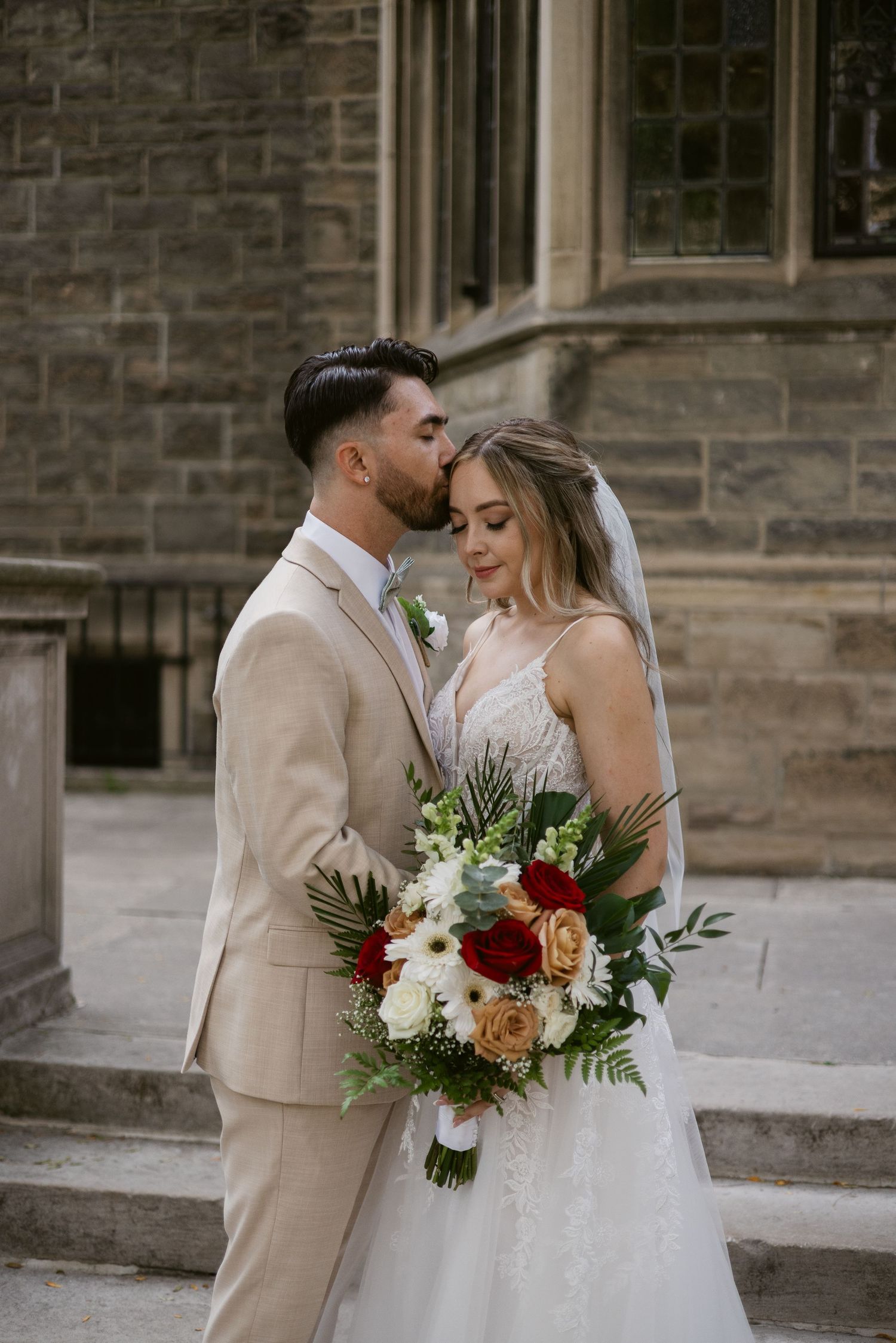 Groom kisses bride's forehead as she holds colorful bouquet of red and peach roses outside stone church.