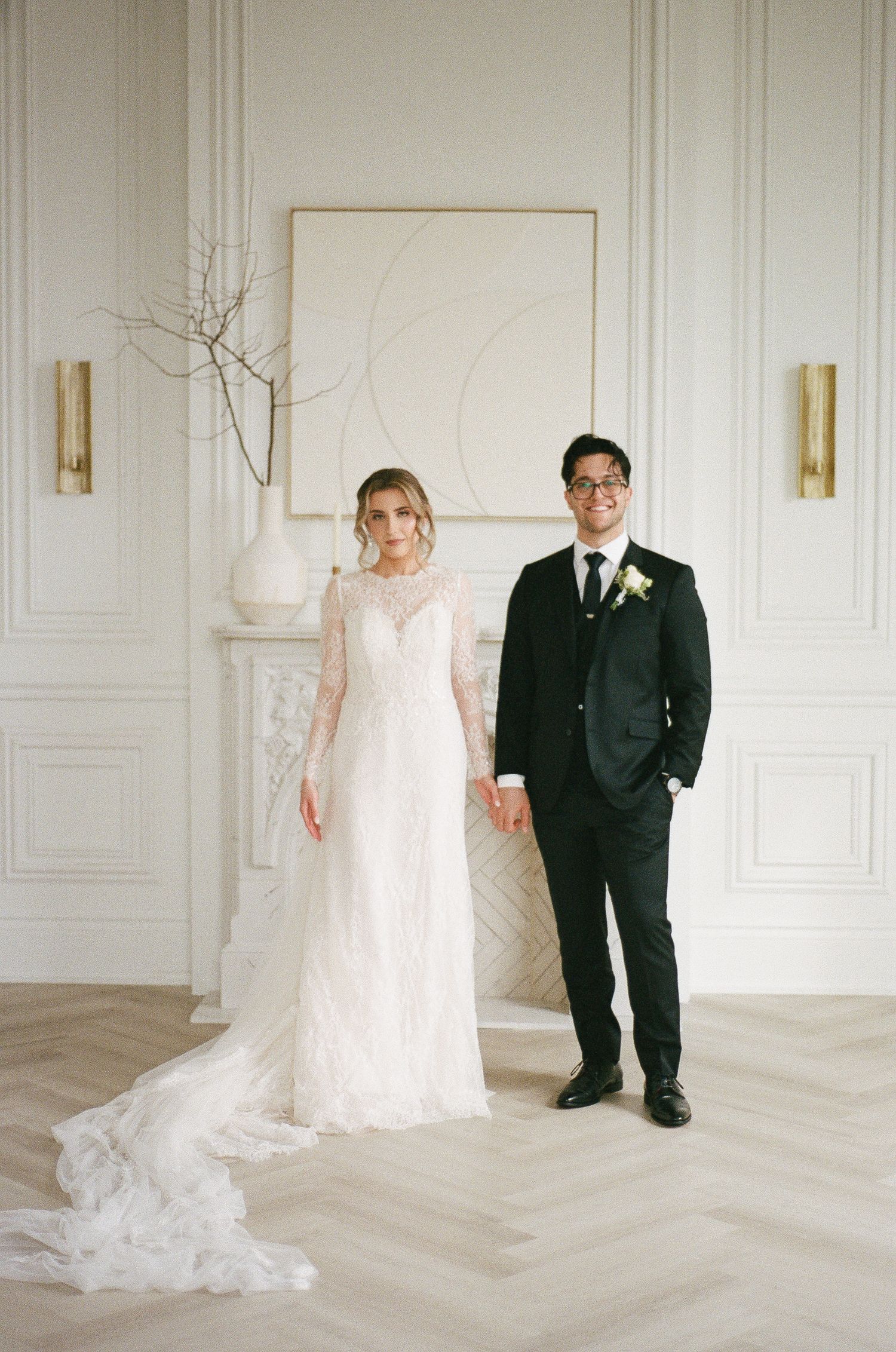 Bride in long-sleeve lace wedding gown and groom in black suit standing together in a bright white elegant room.