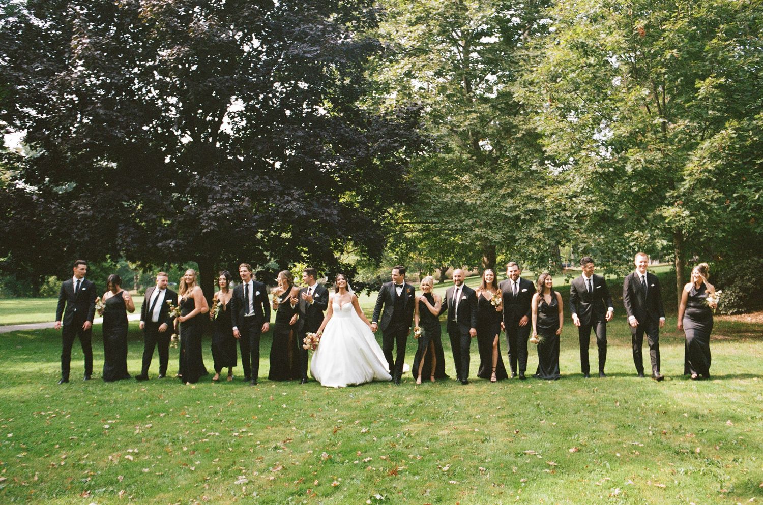 Large wedding party in black attire poses with bride and groom on lush green lawn surrounded by trees.