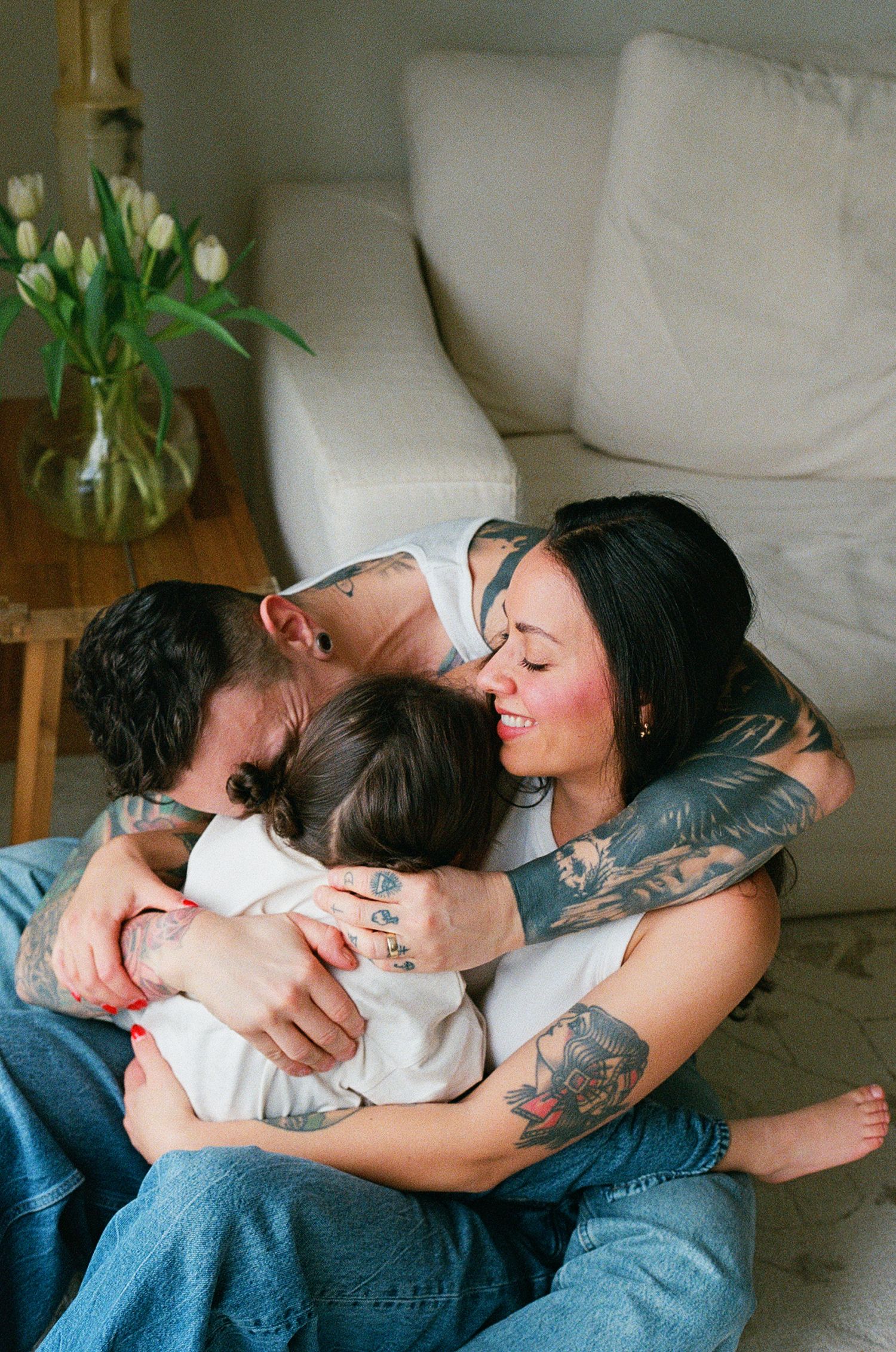 Tattooed couple hugging and kissing their young child on living room floor near white sofa.