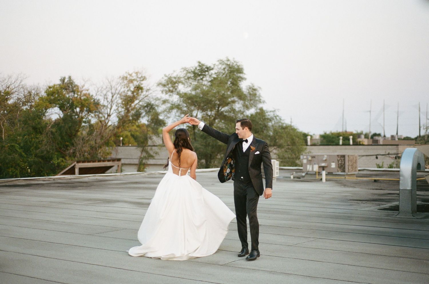 Groom twirls bride in flowing white ball gown on rooftop during joyful wedding photo session.