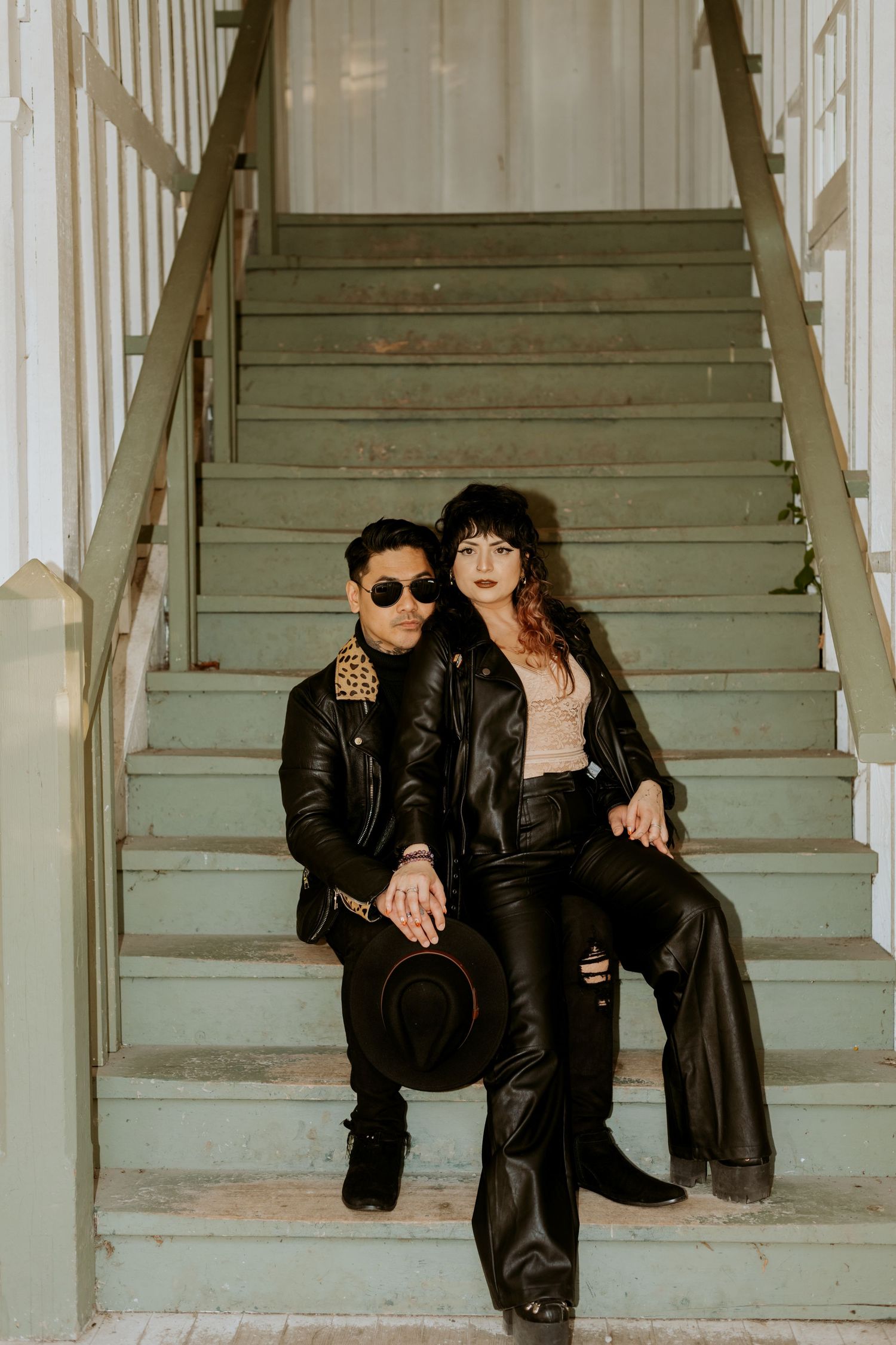 Stylish couple in matching black leather jackets sitting together on vintage green staircase outdoors.