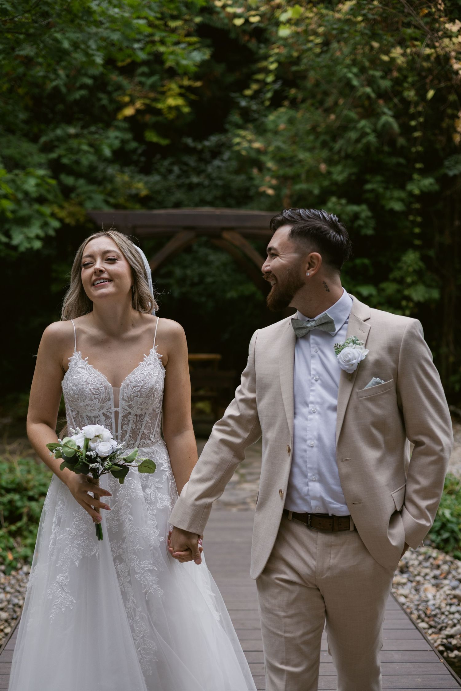 Laughing bride and groom walking together on garden pathway holding hands with white bouquet.