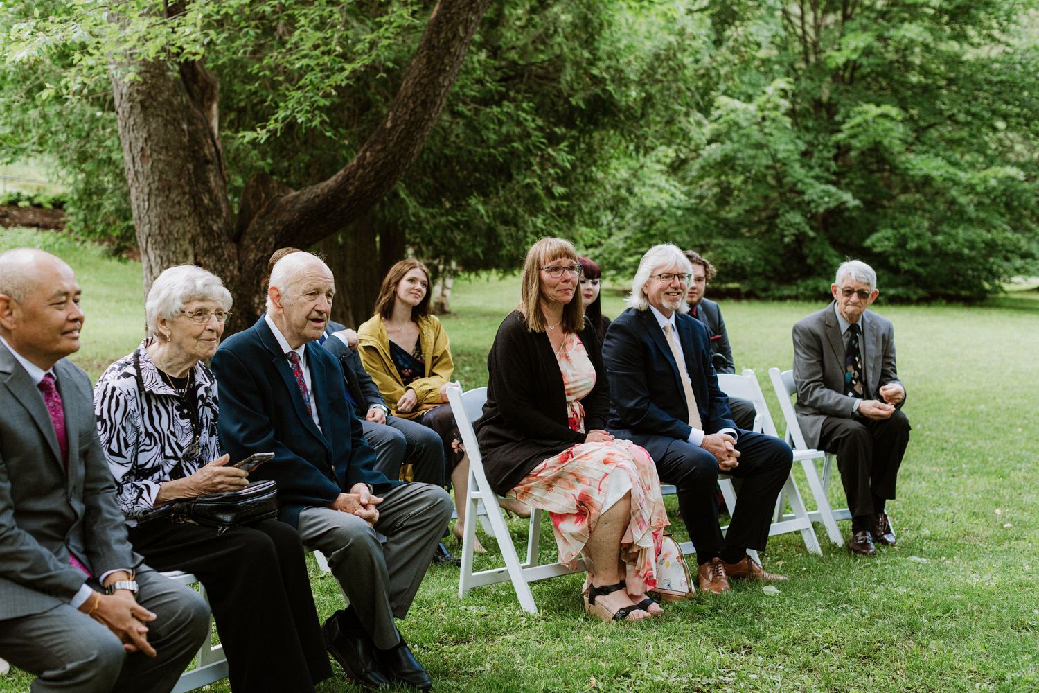 Small group of wedding guests seated on white chairs watching outdoor ceremony surrounded by green trees.