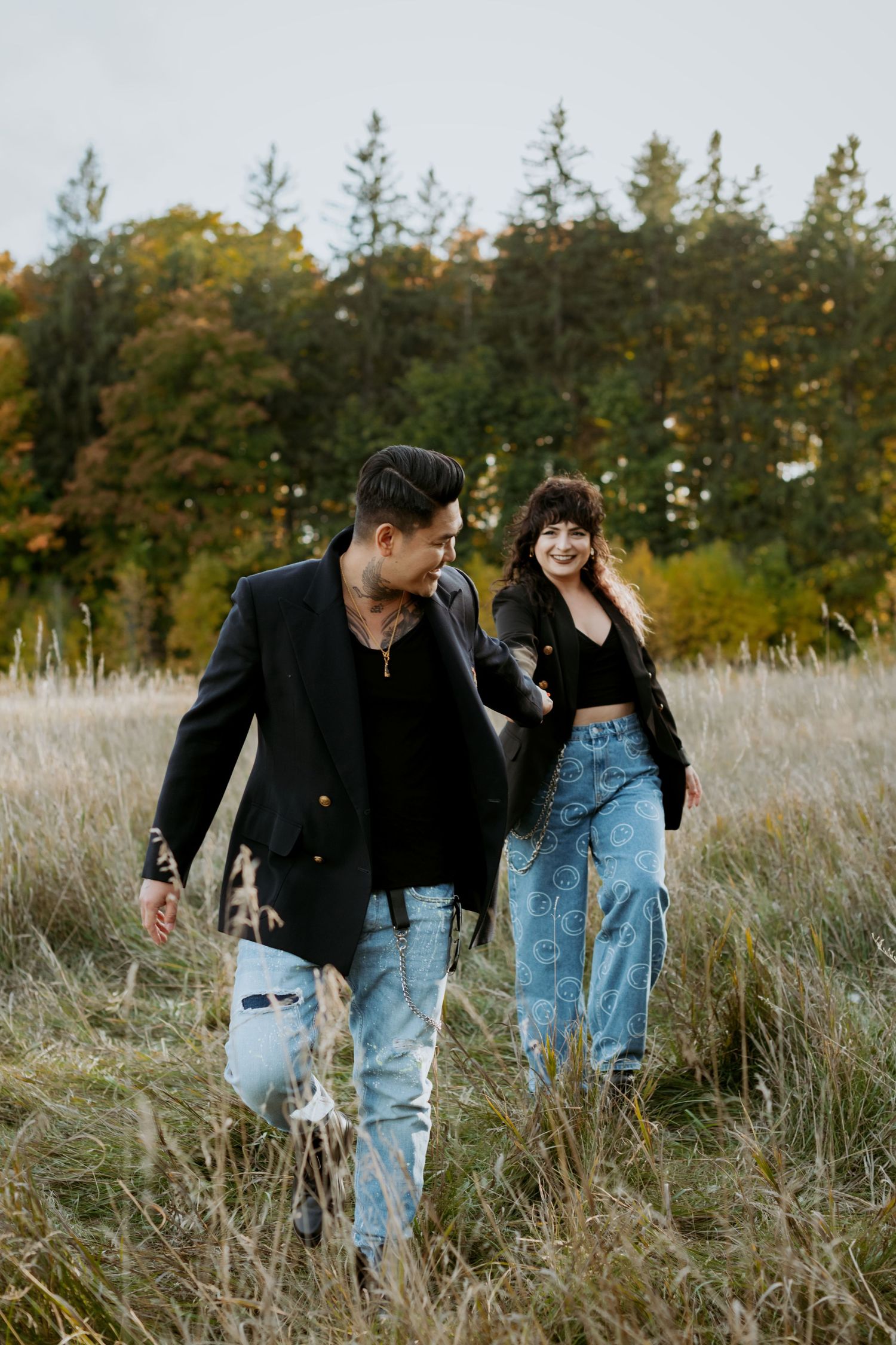 Couple laughing and walking through tall grass meadow in coordinating black blazers and denim jeans in fall setting.