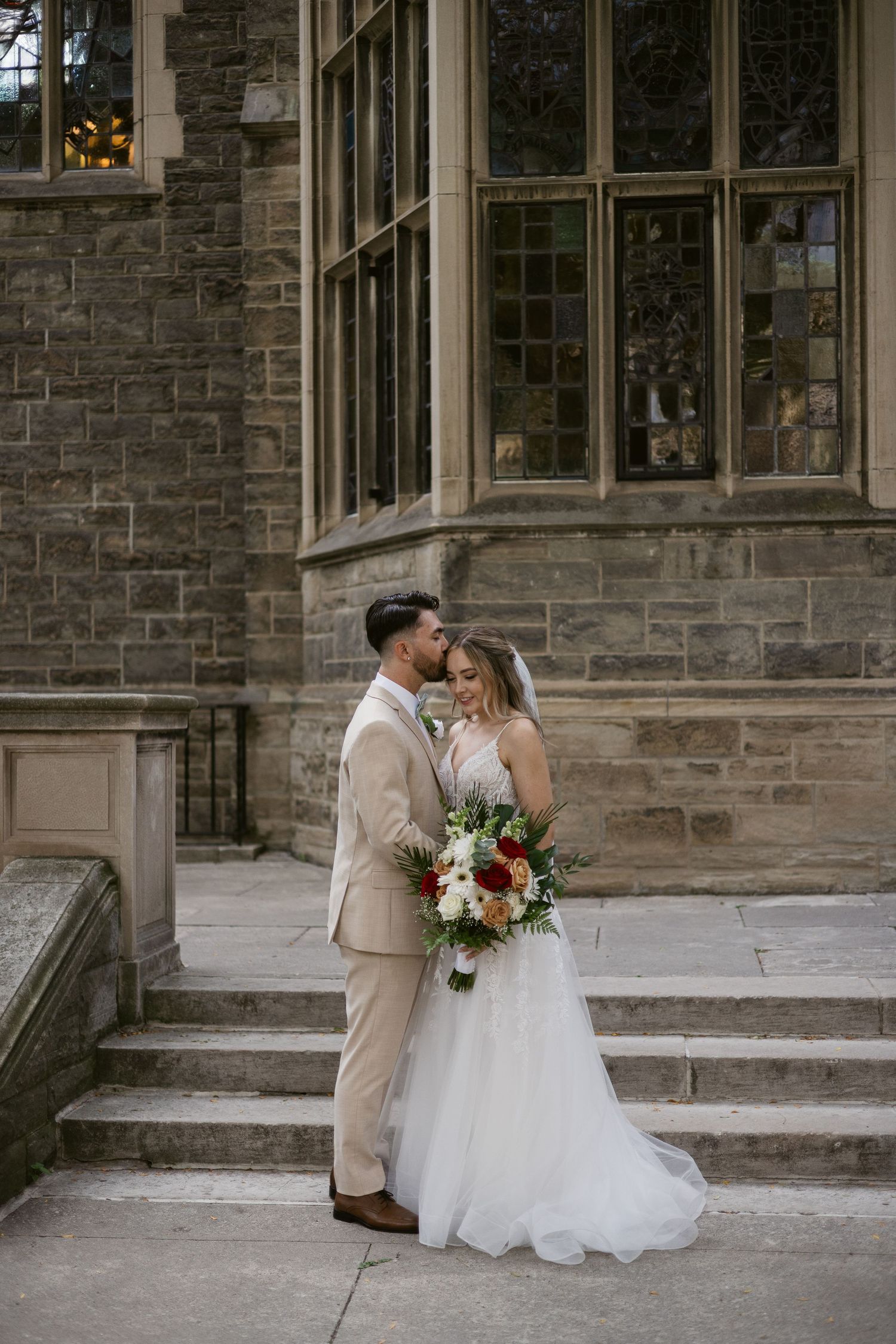 Bride and groom share intimate moment on stone steps holding vibrant floral bouquet at historic venue.