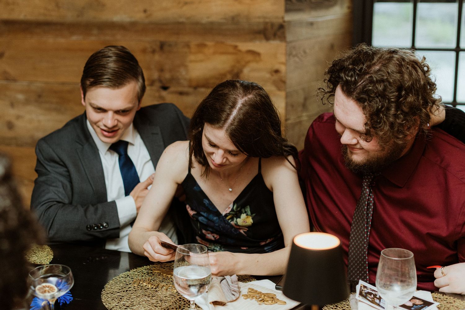 Three wedding guests laugh together while looking at phone at reception dinner table.