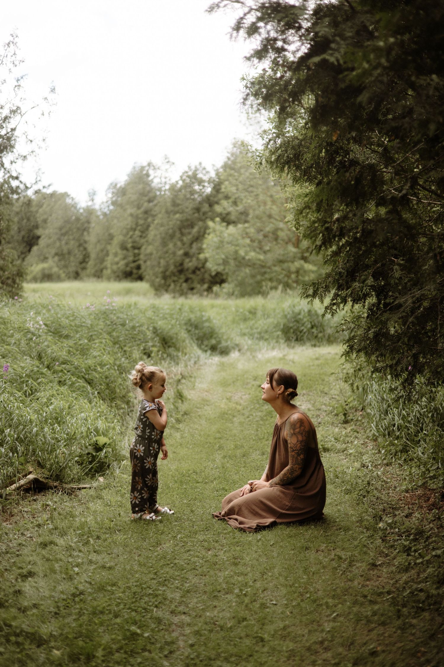 A tattooed mother in a brown dress sits on a garden path facing her young daughter among lush green trees.