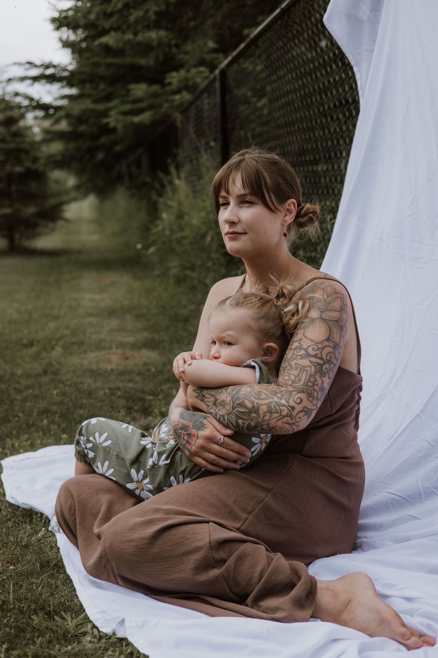 Tattooed mother in brown jumpsuit sits on white blanket outdoors cradling sleeping toddler in floral pants.