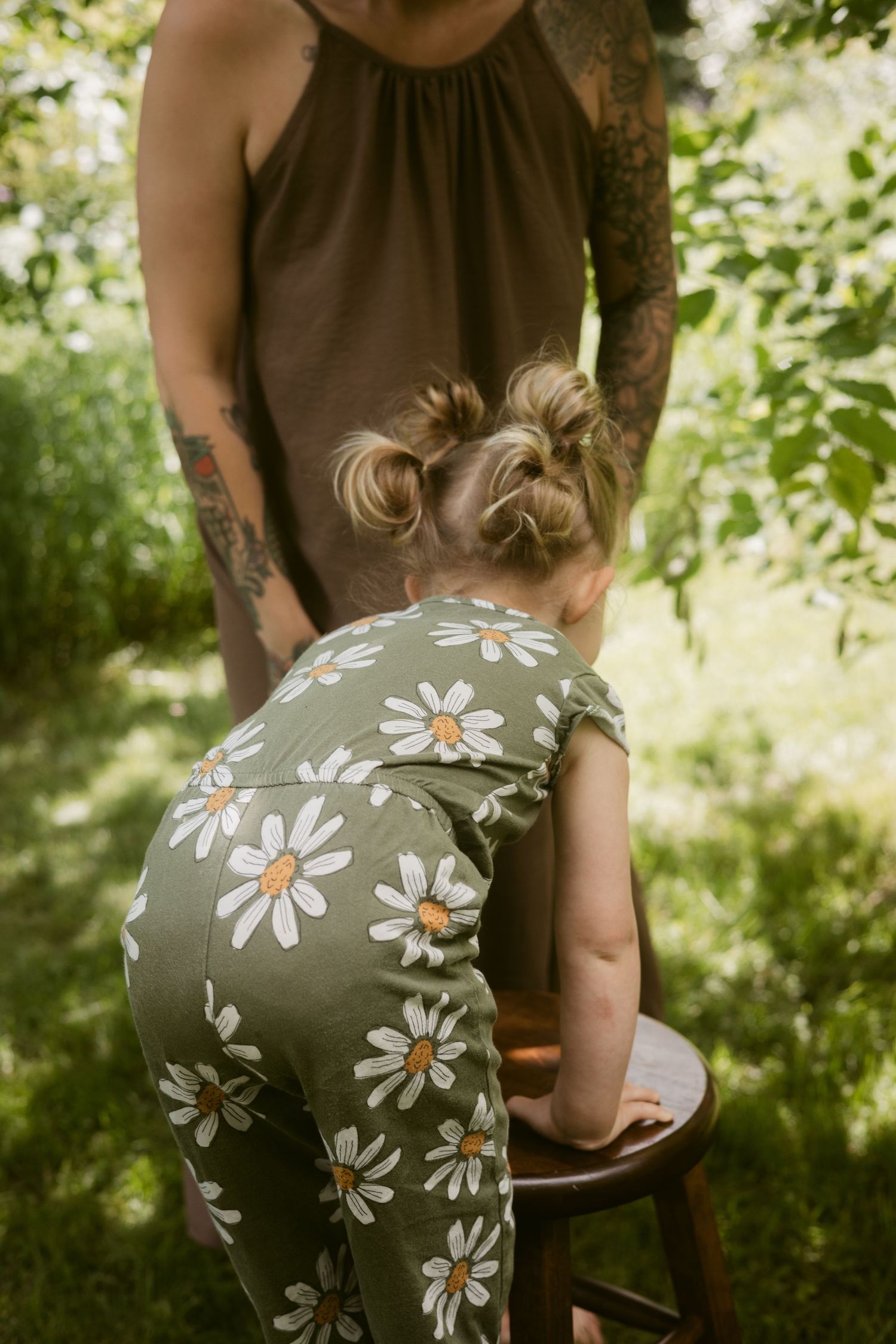 Close-up of a toddler girl in a daisy-print romper leaning over a wooden stool in a garden setting.