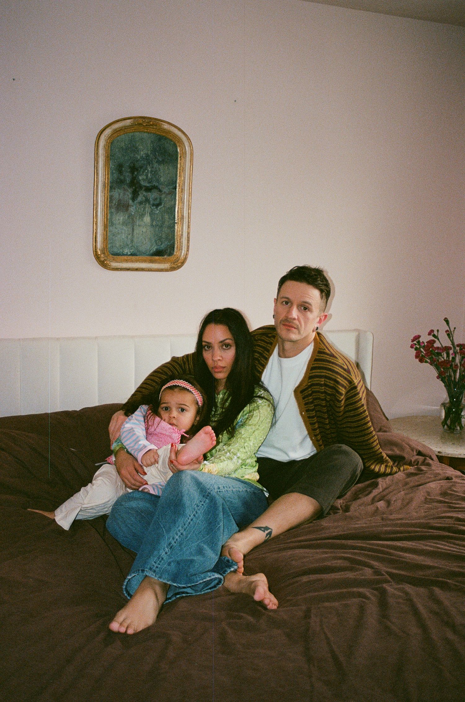Family of three poses on a brown bed, parents holding baby in pink outfit, gold mirror on pink wall behind.