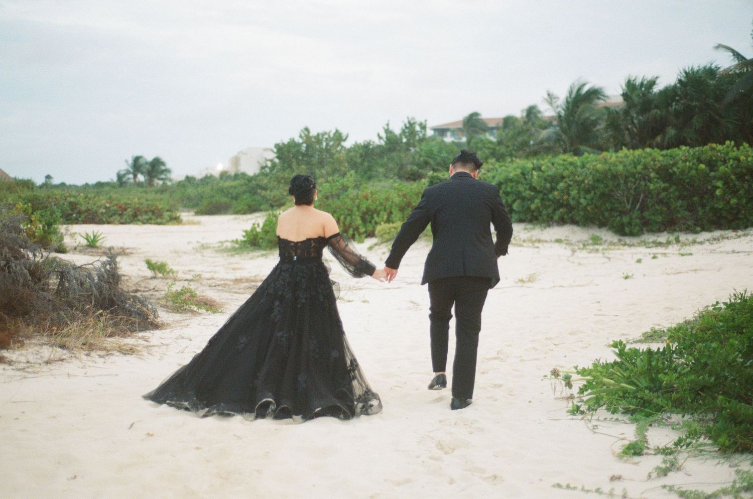 Bride and groom walk hand in hand on white sand beach, bride in flowing black lace wedding gown.