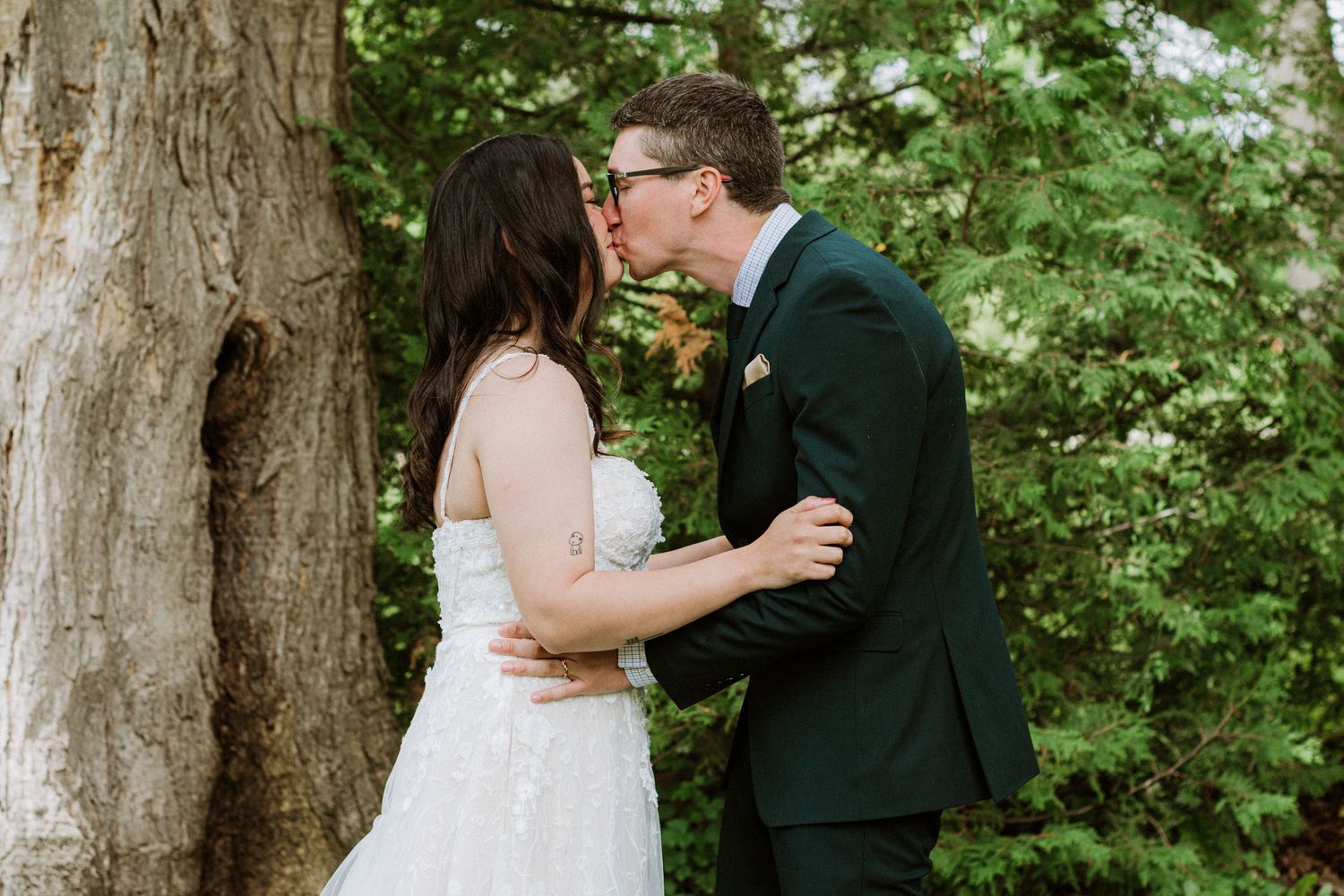 Bride and groom sharing first kiss surrounded by green trees at outdoor wedding ceremony.