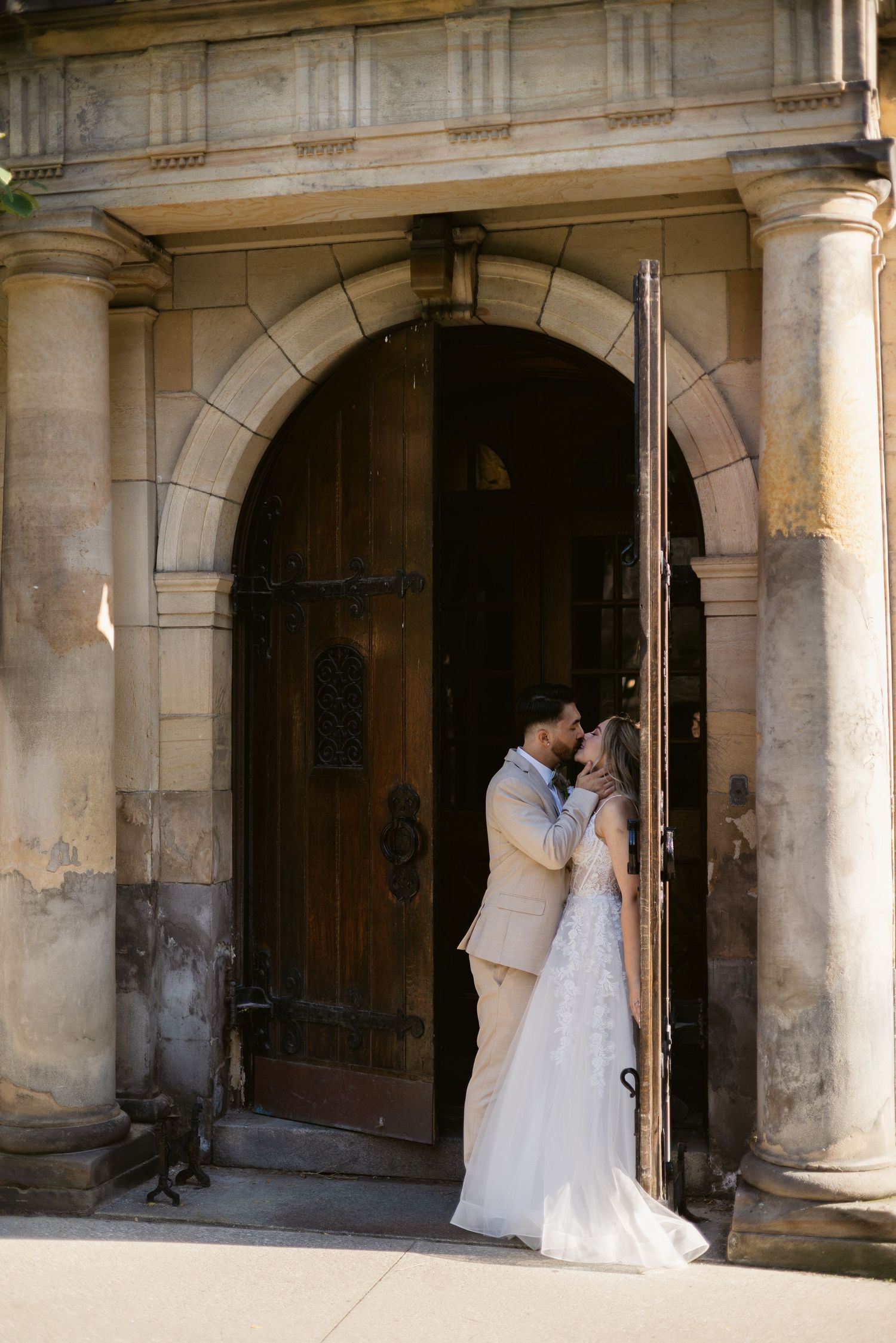 Wedding couple kisses in front of grand arched wooden doors of an elegant stone building.