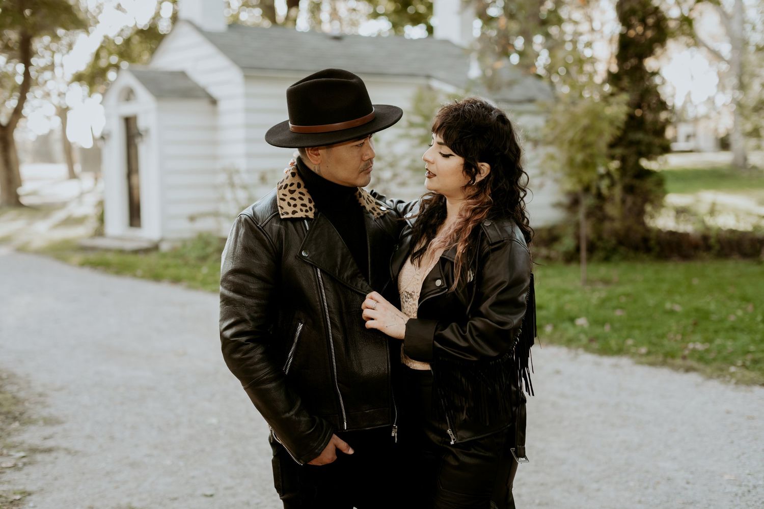 Couple in leather jackets and wide-brim hat sharing an intimate moment on a tree-lined gravel path.