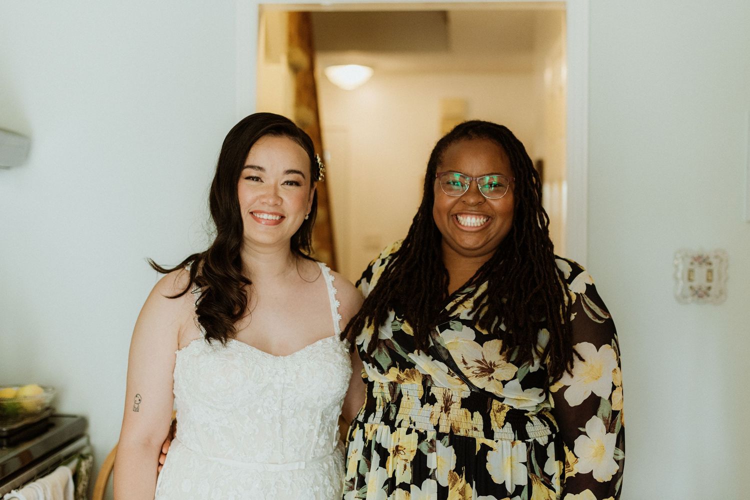 Bride in white lace gown smiling with wedding planner in floral dress during bridal preparations.