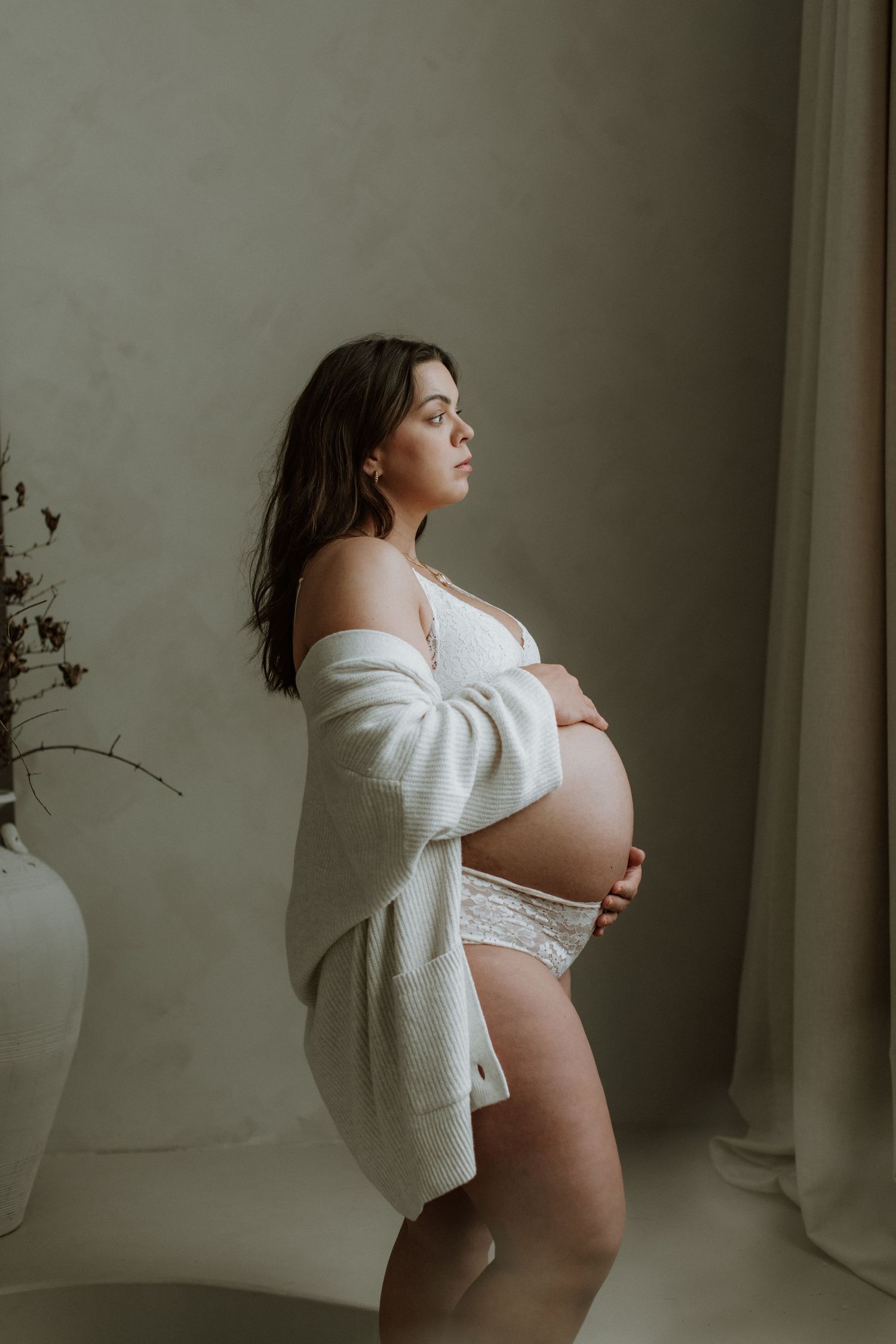 Pregnant woman in white lace lingerie and cardigan cradling her baby bump near a window.