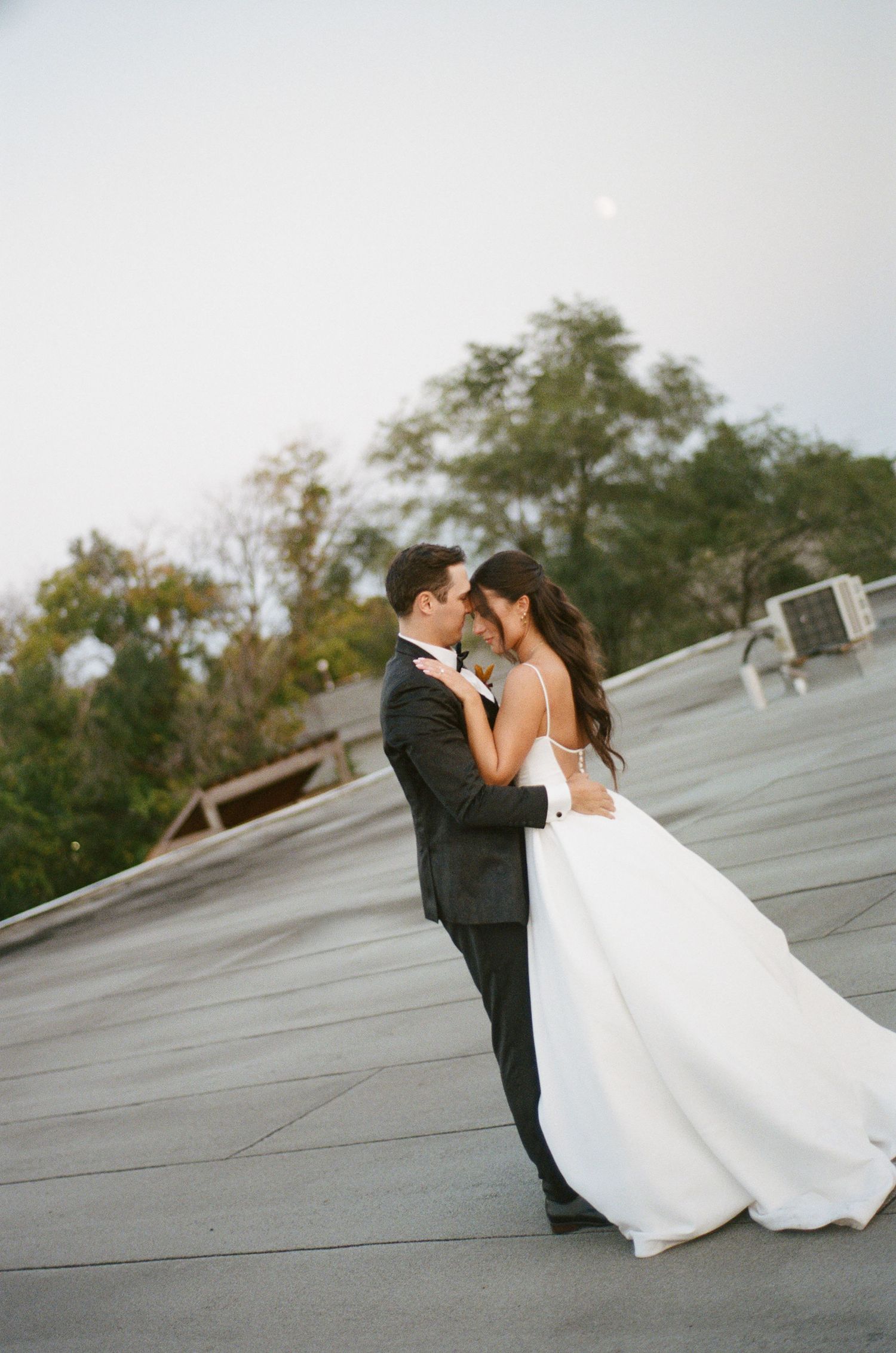 Bride in elegant white ball gown and groom in tuxedo sharing a romantic embrace on a rooftop.