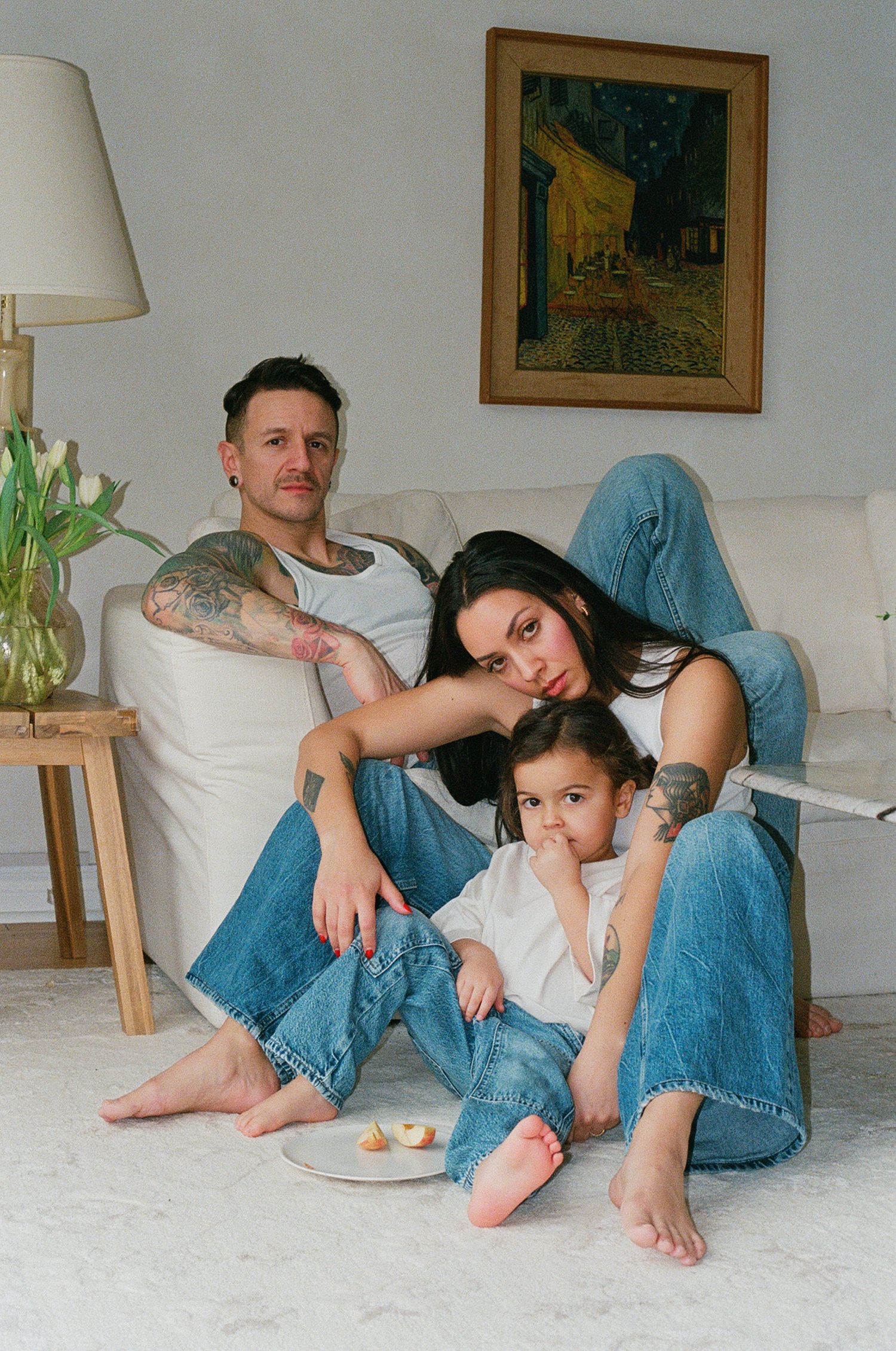 A tattooed man, a woman and a young child in matching blue jeans relax barefoot on a white rug beside a sofa with a snack plate.