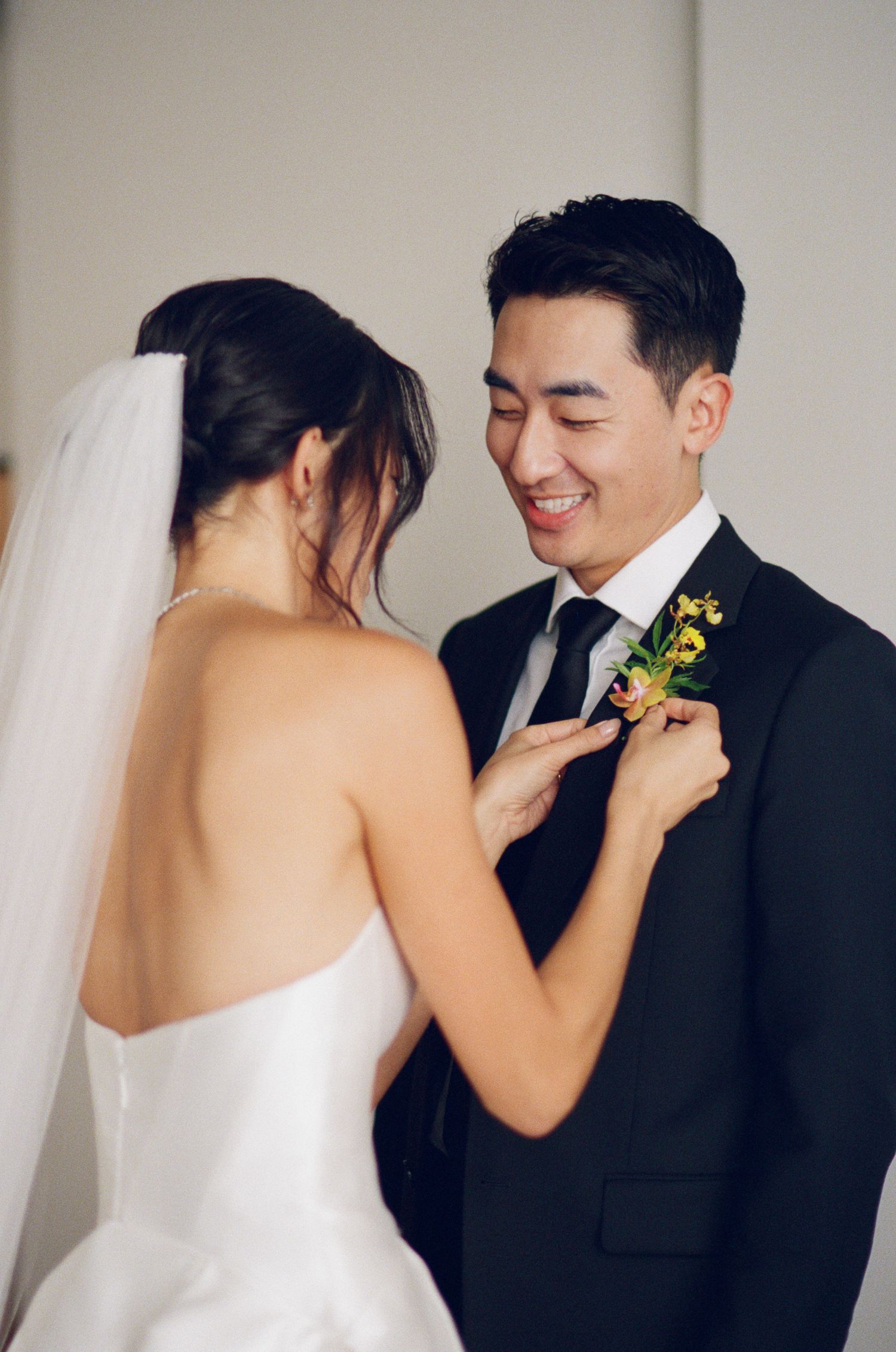 A smiling groom in a black suit watches as his bride pins a yellow boutonniere to his lapel.