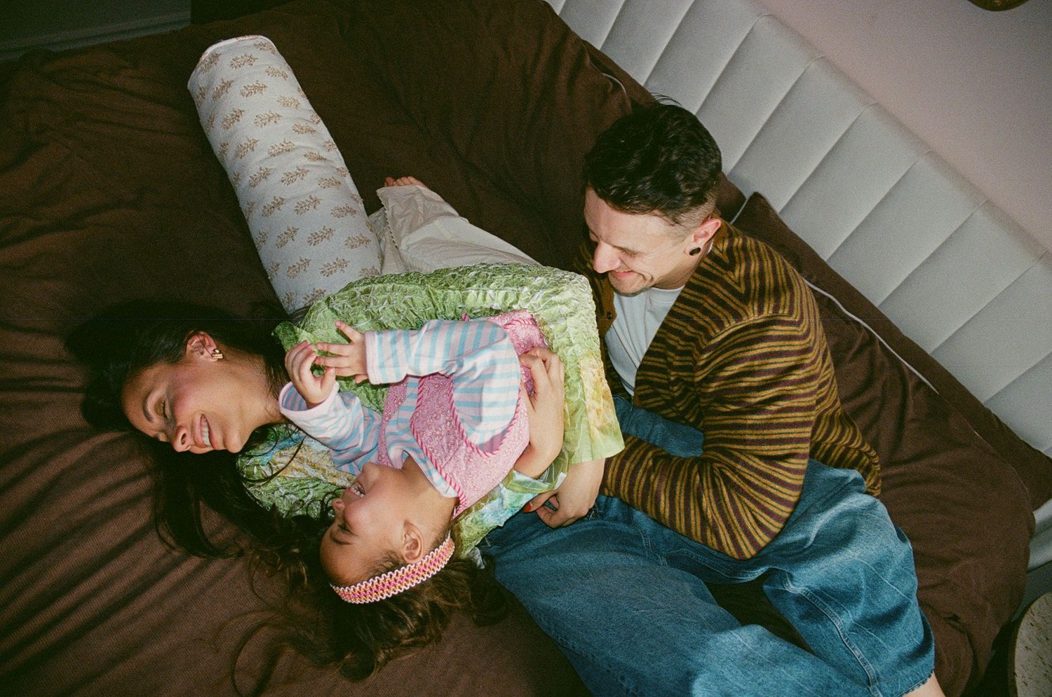 A happy family laughs together while lying on a brown sofa, captured from above in a warm indoor setting.