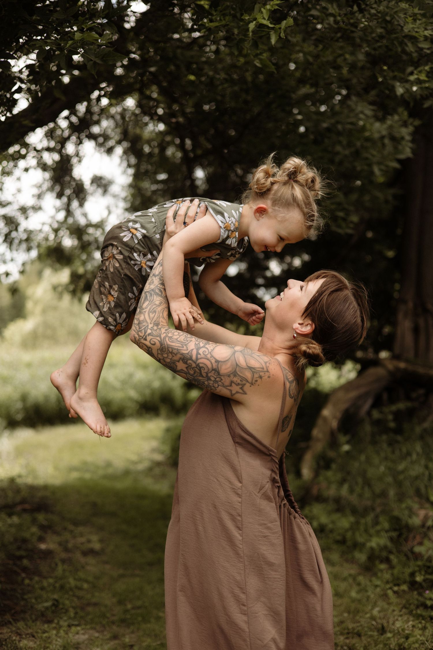 Tattooed mother in brown dress lifts smiling toddler in the air in a green garden.