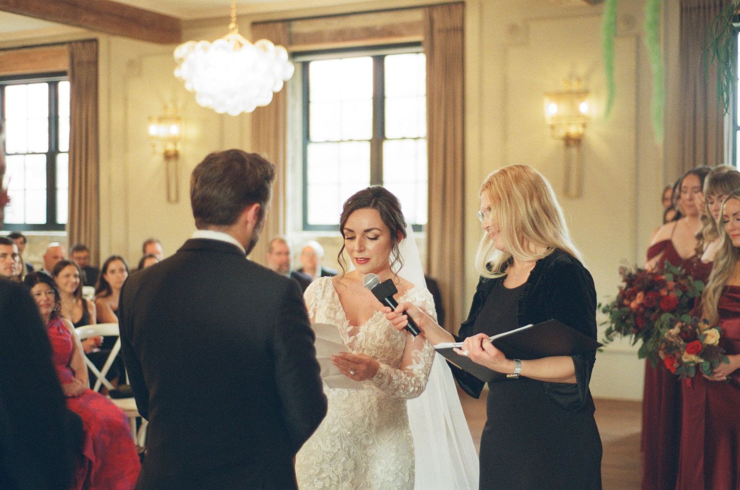 Bride reading vows to groom during indoor wedding ceremony with guests seated behind them.
