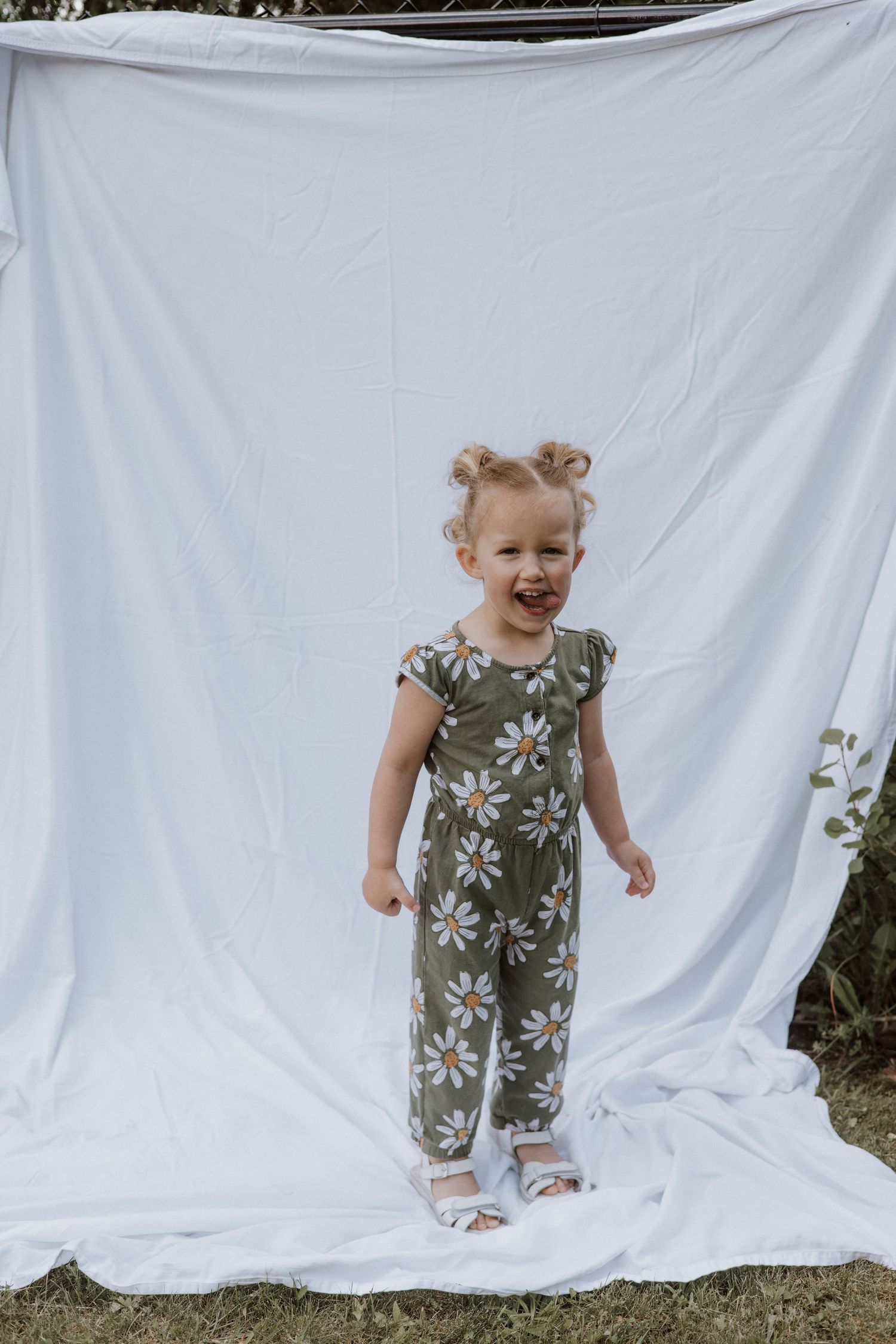 A cheerful toddler girl in a green daisy print jumpsuit laughs while standing in front of a white fabric backdrop outside.