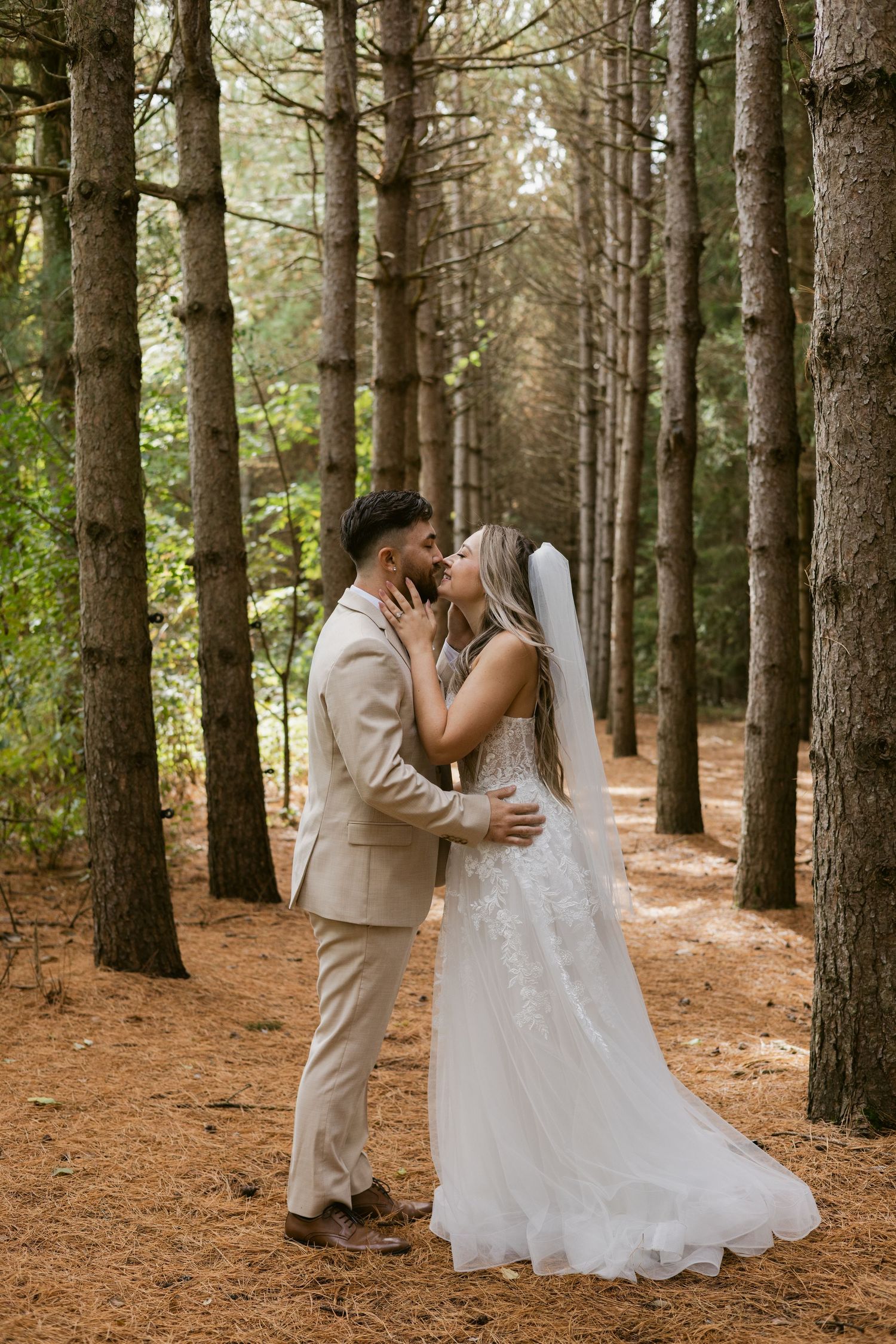 Bride and groom share a romantic kiss among tall pine trees, dressed in wedding attire in a forest setting.