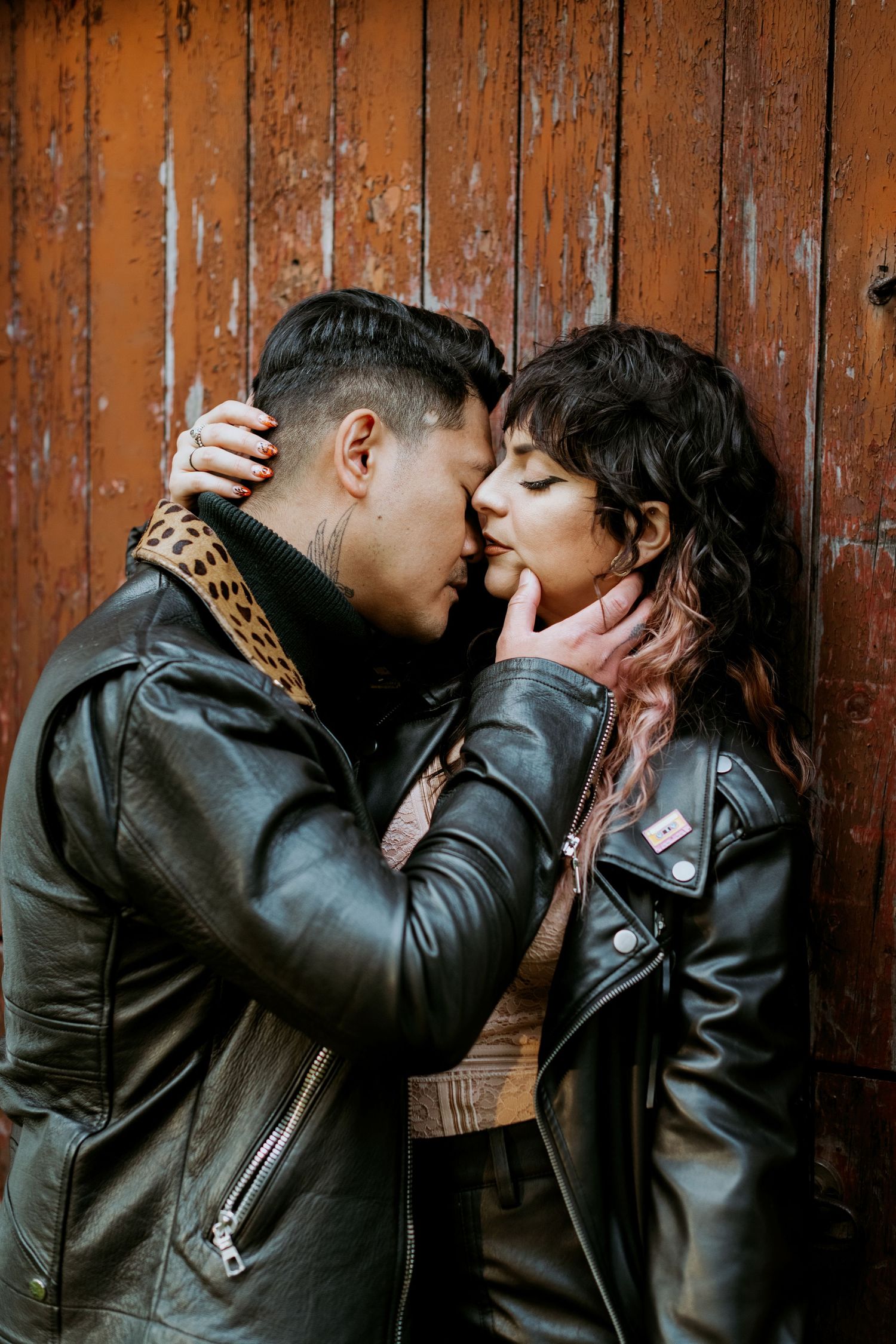 Couple in matching leather jackets sharing intimate moment against rustic wooden door.