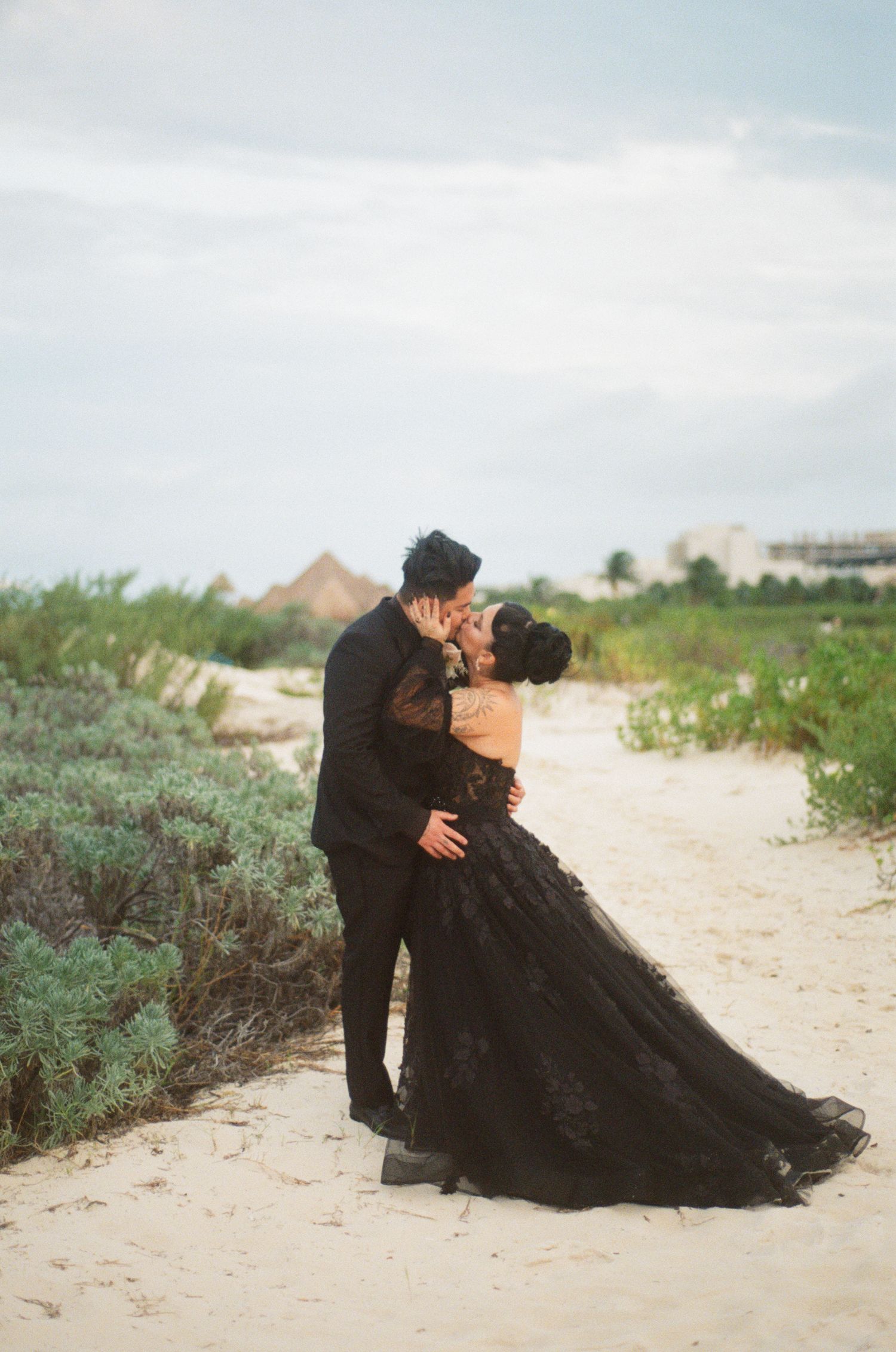 Couple kissing on sandy beach path, bride in black lace ballgown, groom in black suit, coastal greenery background.
