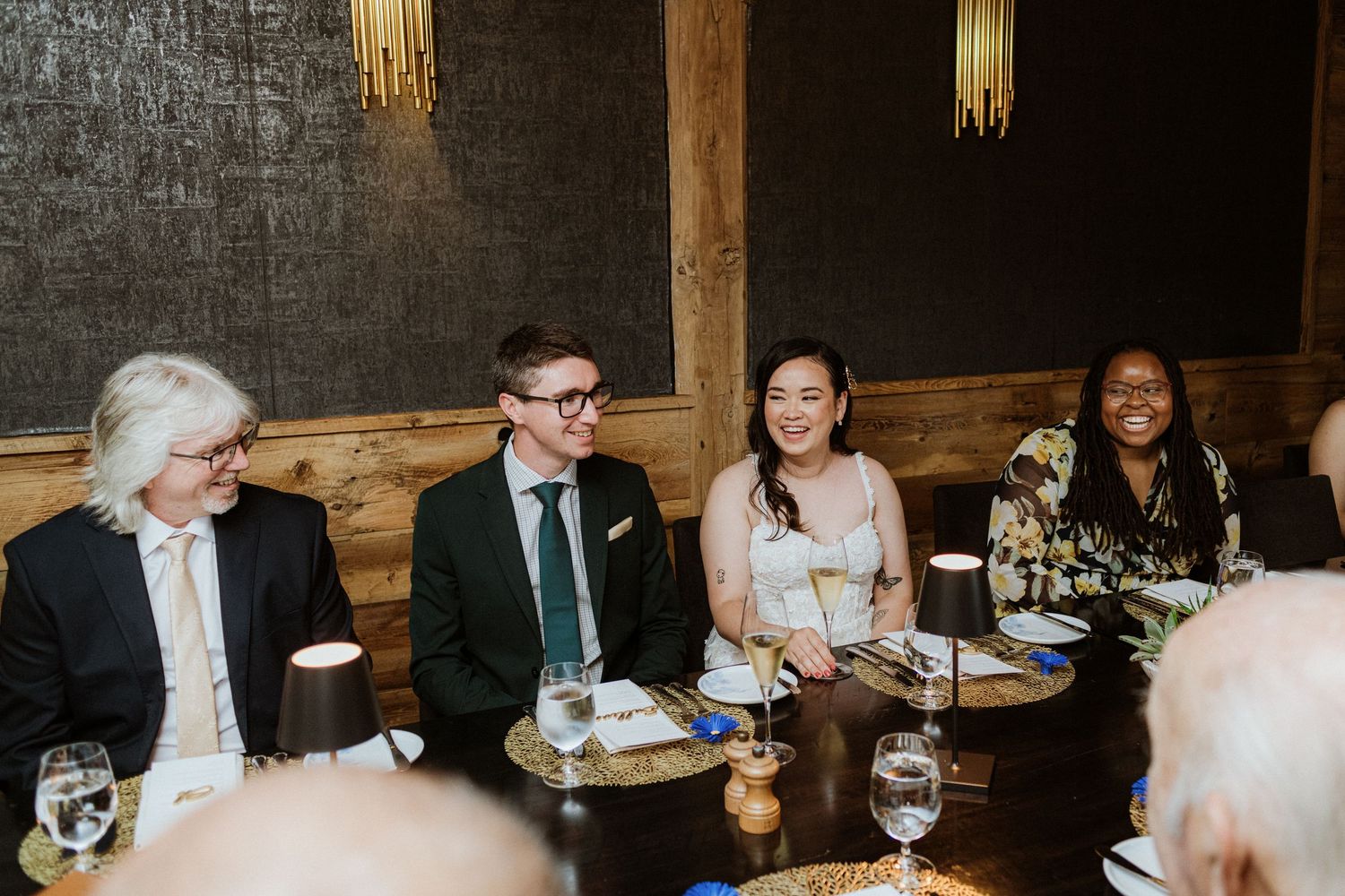 Wedding guests smiling and laughing together at a reception dinner table in a rustic restaurant.