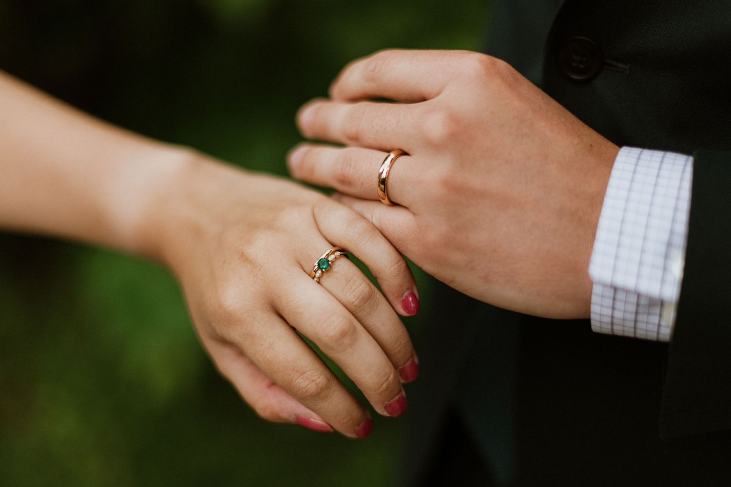 Close-up of bride and groom holding hands showing emerald and gold wedding rings outdoors.