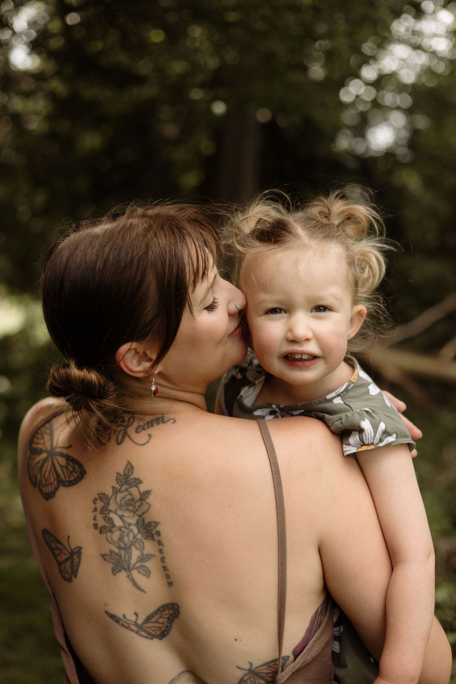 A tattooed mother with butterfly back tattoos kisses her laughing toddler daughter outdoors.