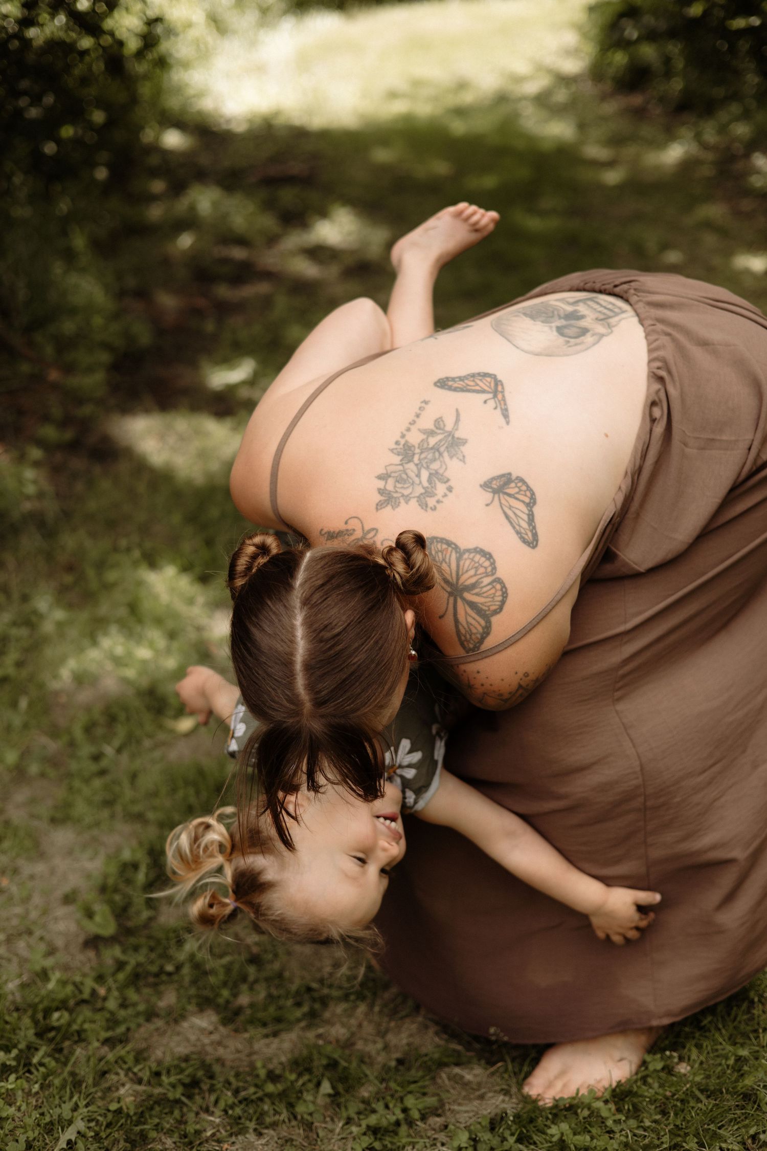 A tattooed mother bends down to kiss her barefoot toddler on green grass.