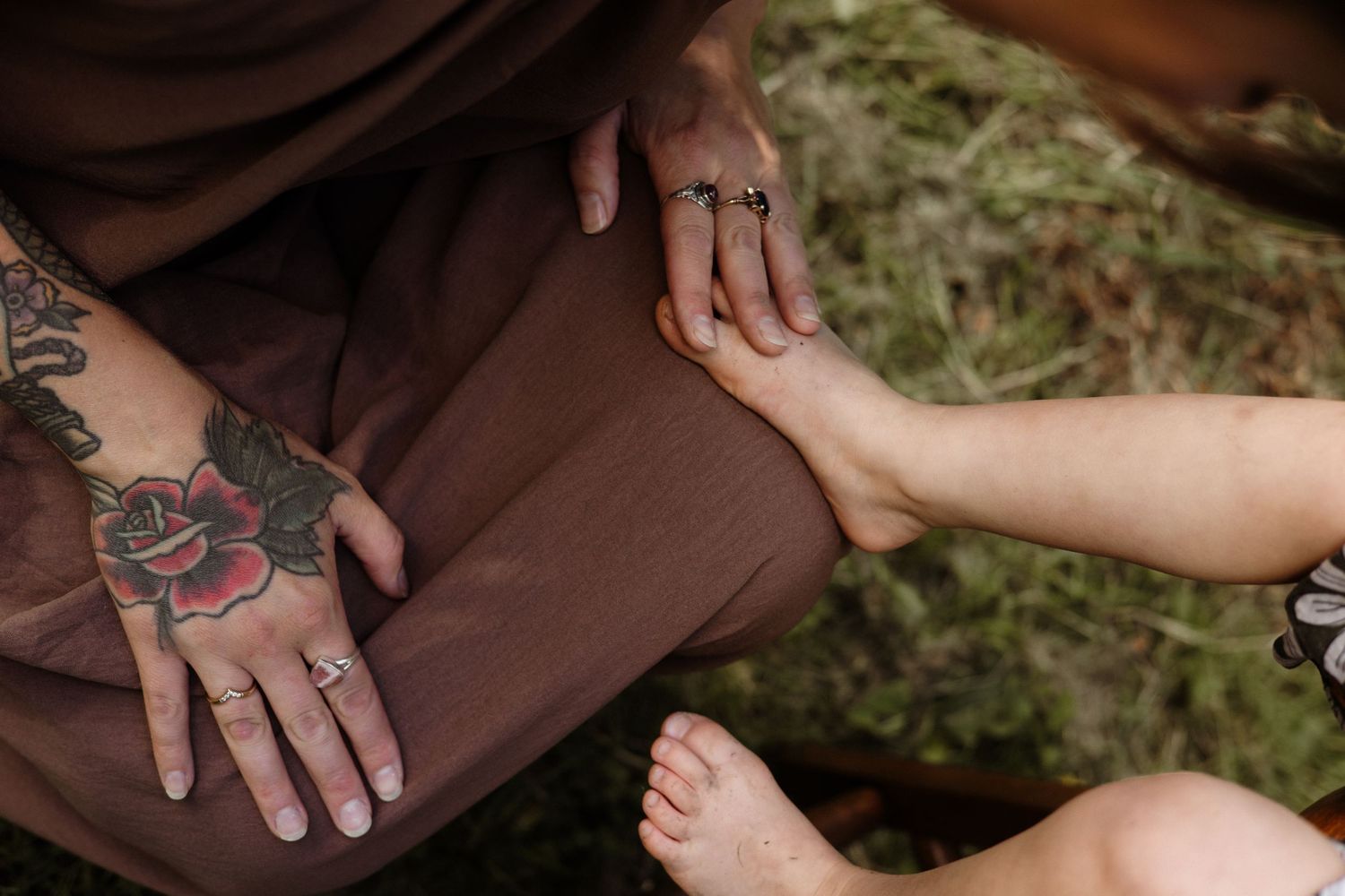 Close-up of tattooed hand with rose tattoo and rings gently holding a small childs bare foot outdoors.