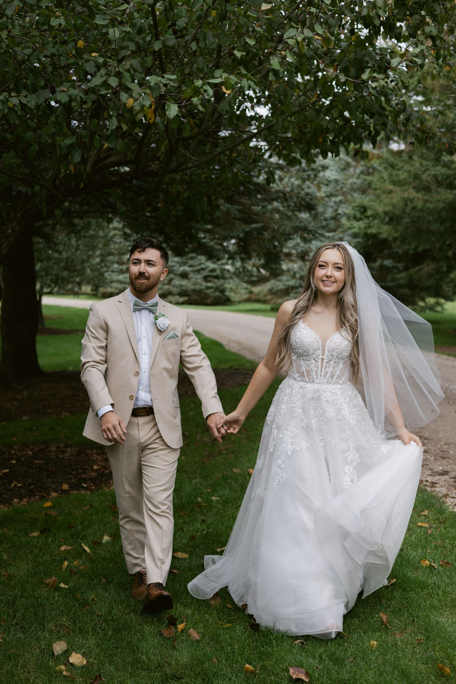 Bride and groom walk hand in hand on green grass, she in a flowing lace gown with long veil.
