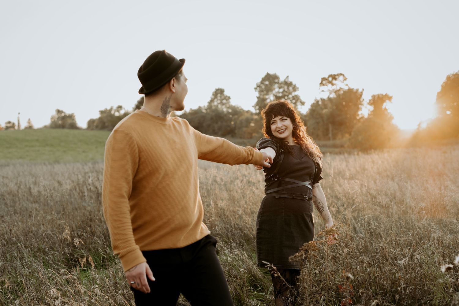 Man in mustard sweater holding womans hand while she smiles back at him in a sunlit golden field.