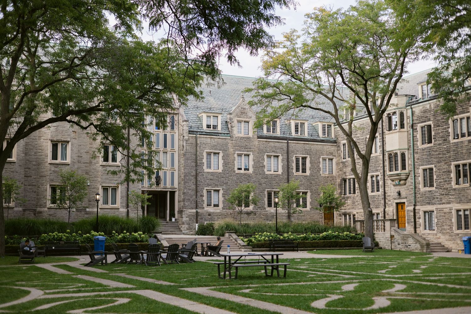 Wide view of a Gothic stone university courtyard with patterned lawn, mature trees, and outdoor seating area.