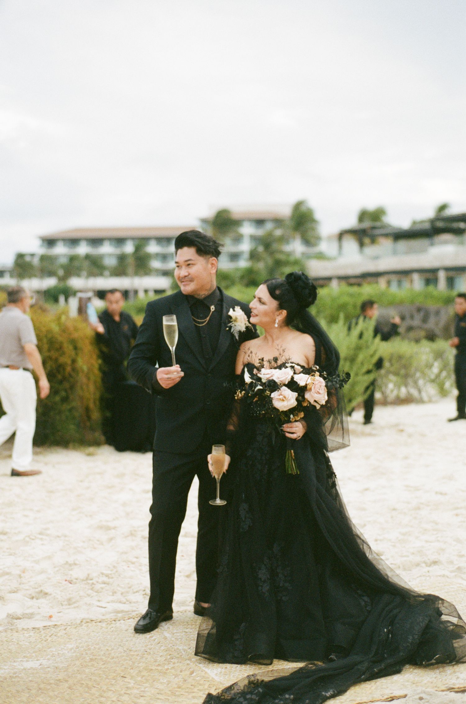 Newlyweds toasting champagne on beach, bride holding floral bouquet in black gown, resort background.