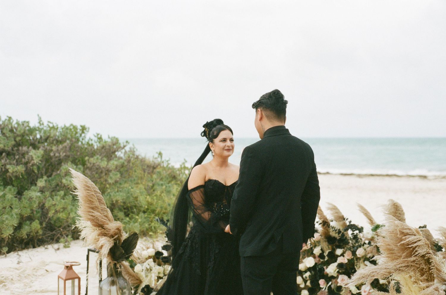 Bride in black wedding gown and groom in black suit exchanging vows at a beach ceremony with pampas grass decor.