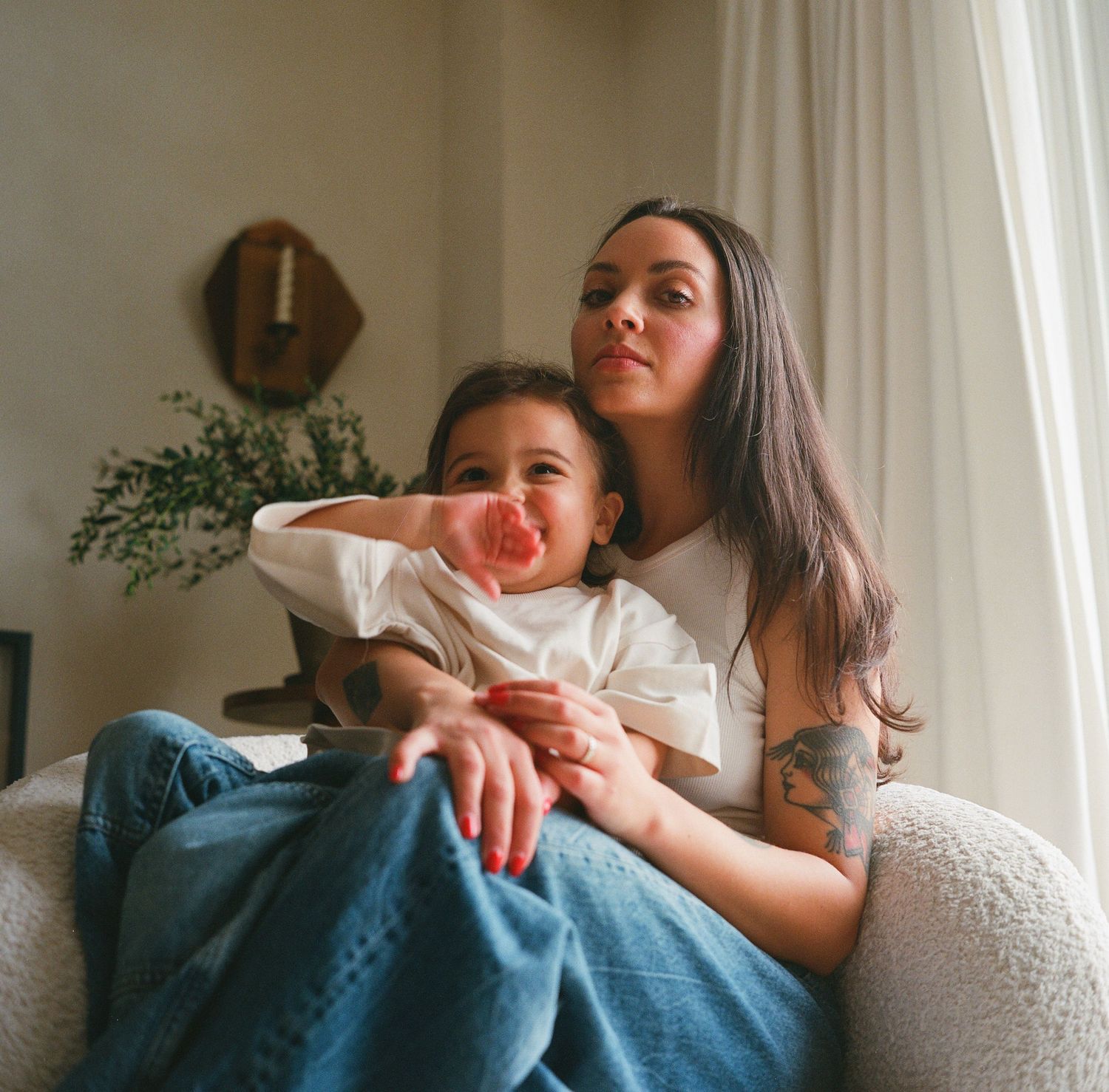 A woman with tattoos sits in a white chair by a window, holding a young child eating a pink treat.