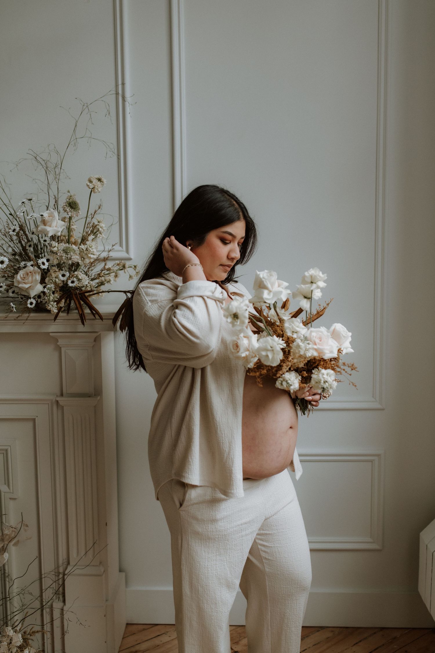 Pregnant woman in beige outfit holding white floral bouquet in elegant room.