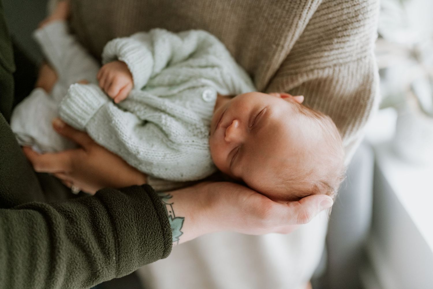 Parent tenderly cradling sleeping newborn in mint knit outfit near a bright window.