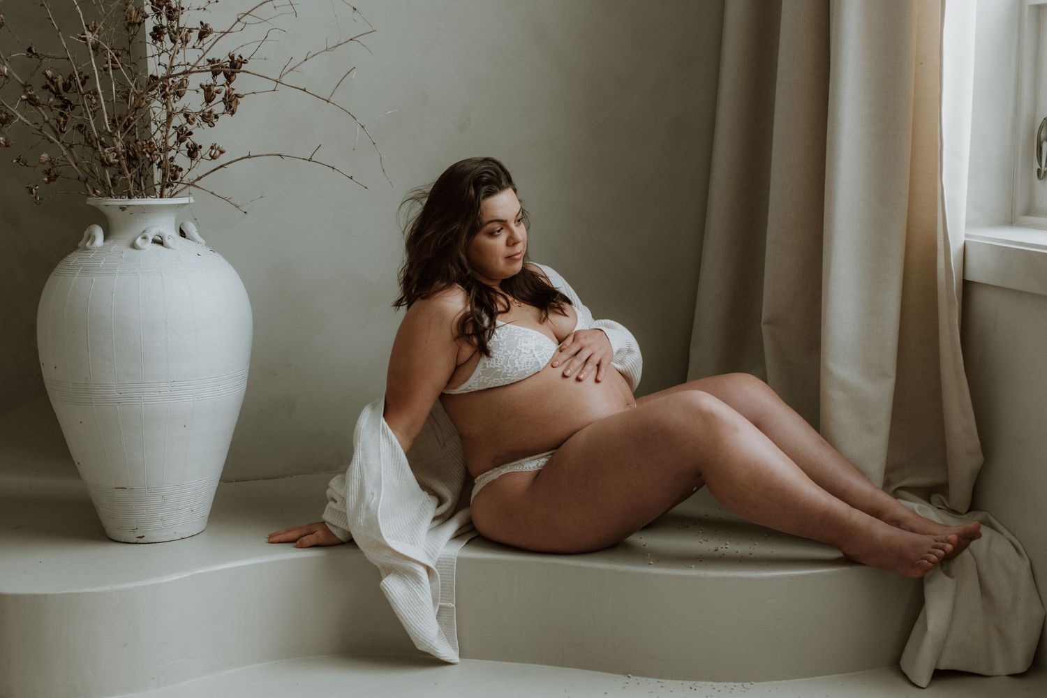 Pregnant woman in white lingerie reclining elegantly beside large white vase near sunlit window.