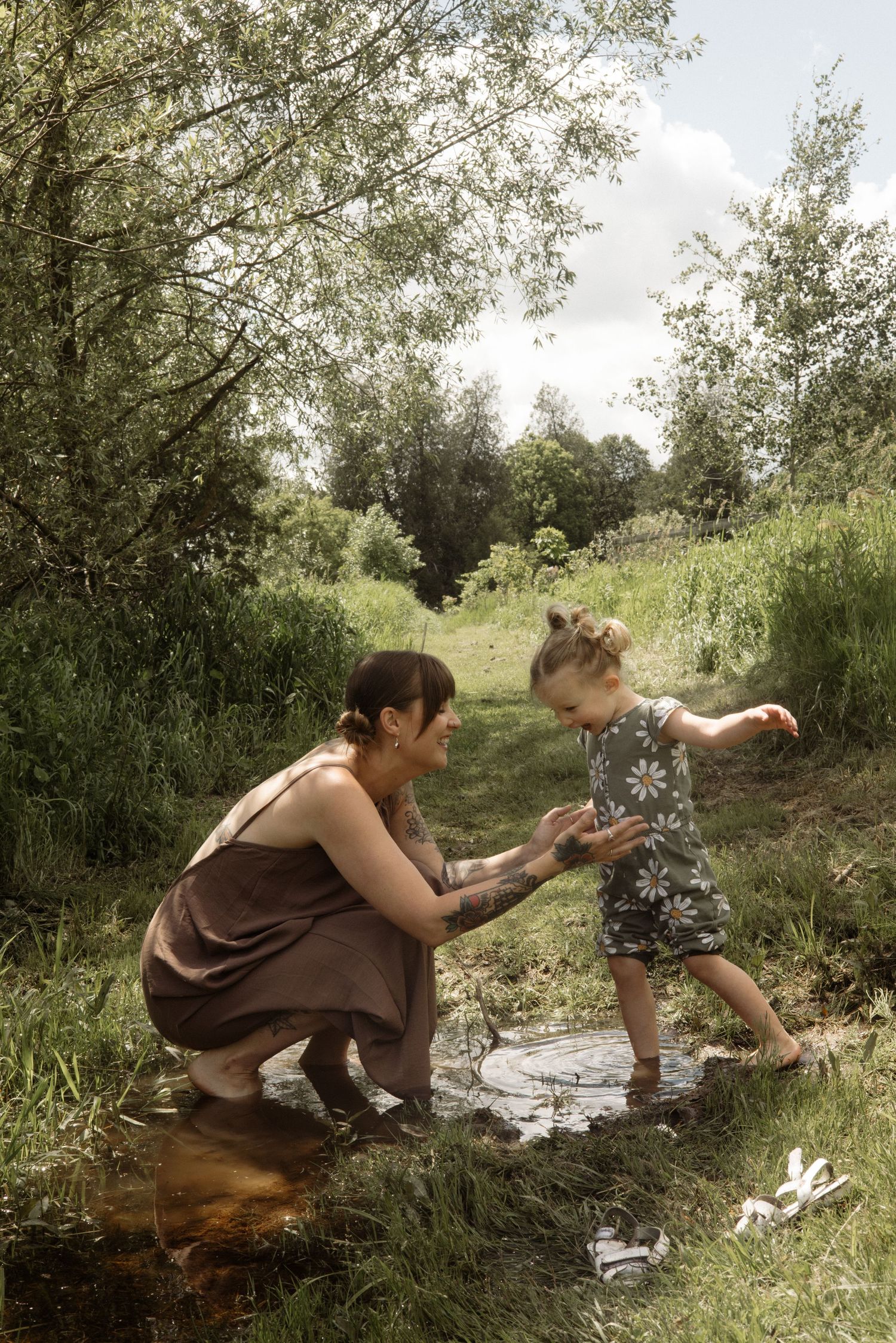 Mother with tattoos kneeling in a green meadow holding her toddler girl's hands near a small puddle outdoors.