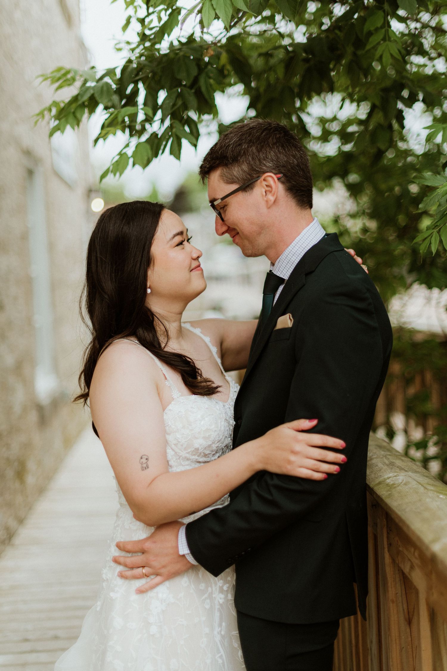 Bride and groom embracing under green tree leaves in outdoor wedding portrait.