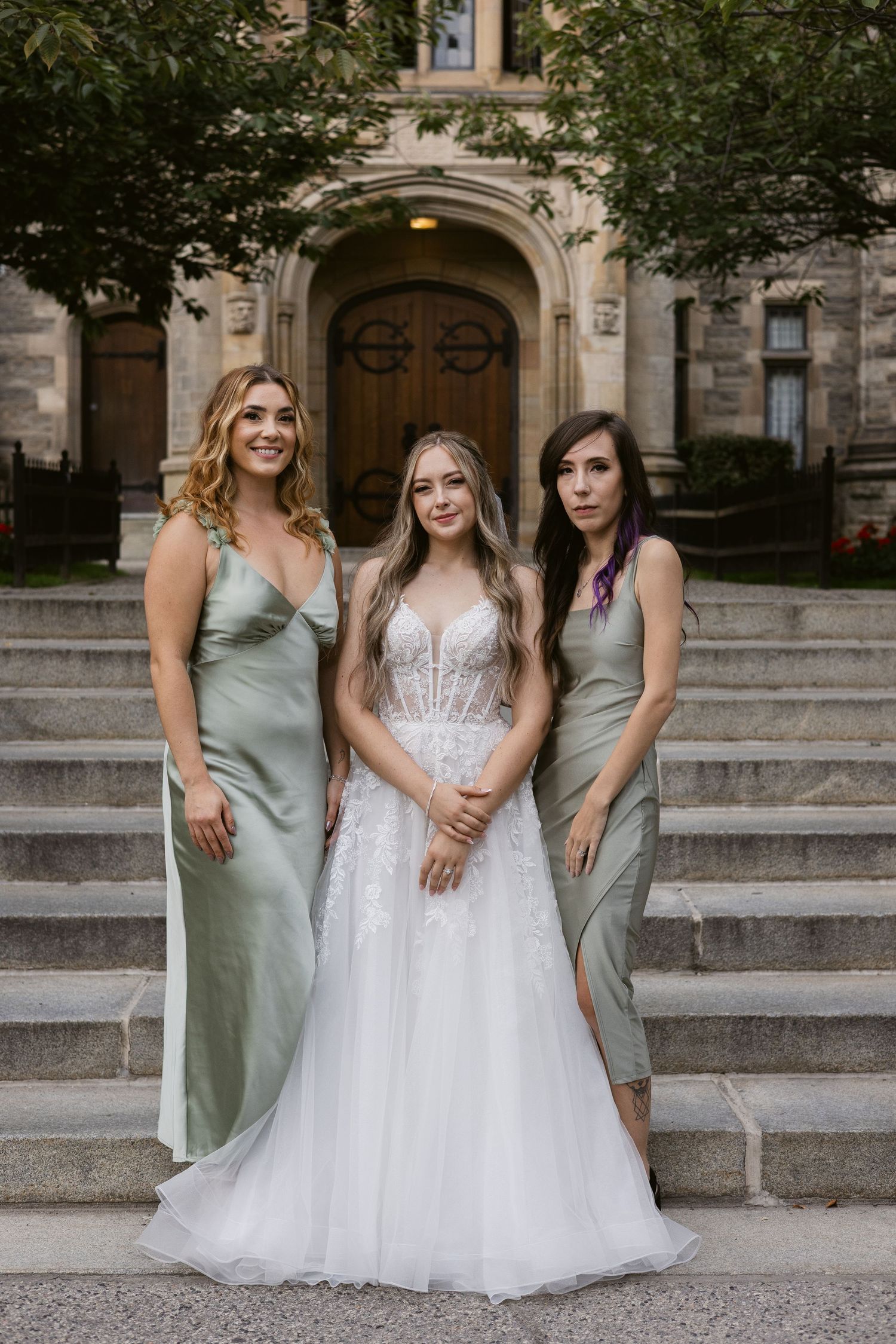 Bride in lace gown poses with two bridesmaids in sage green satin dresses on stone building steps.