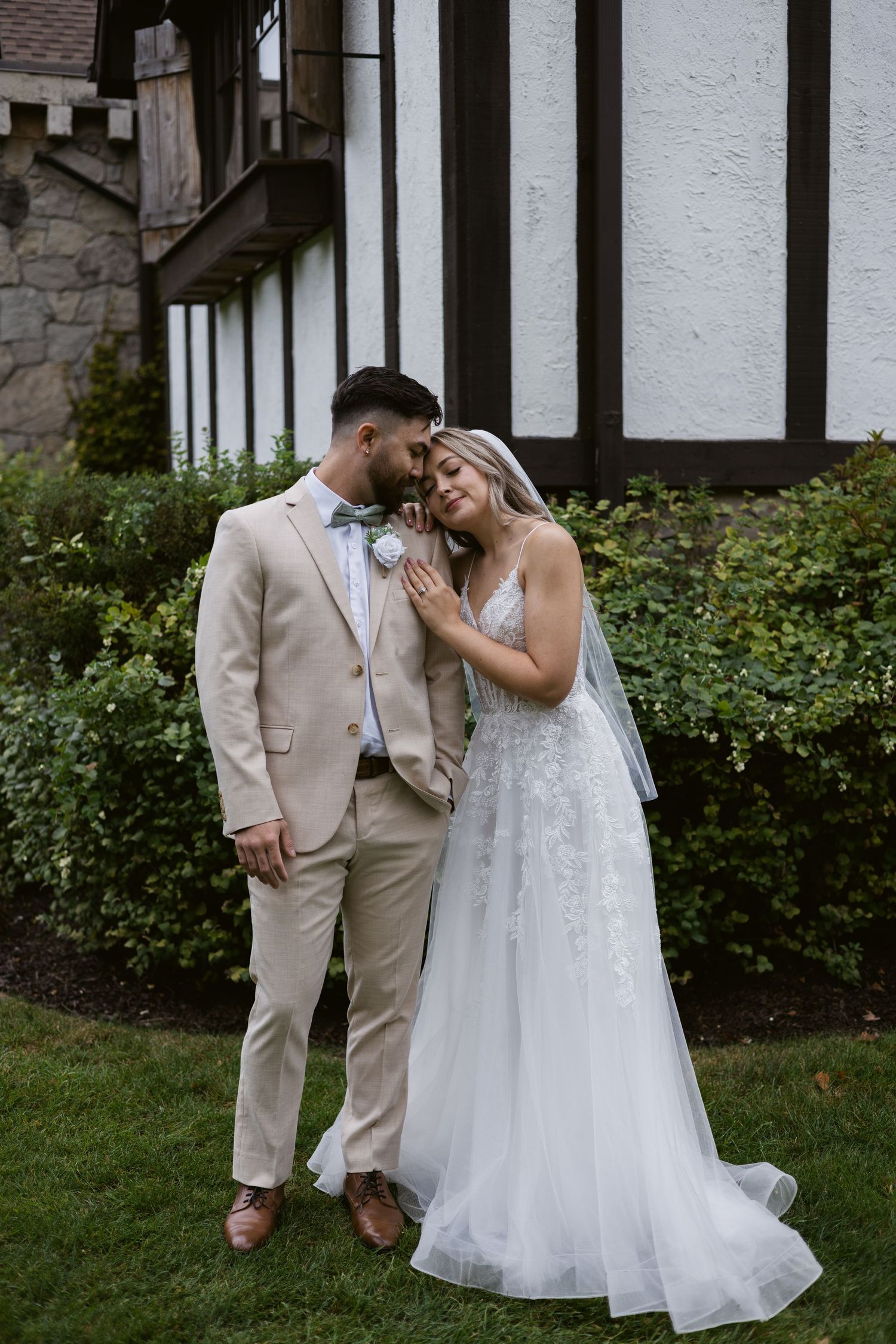 Bride and groom sharing a romantic moment on green lawn, bride in flowing lace gown with cathedral veil.